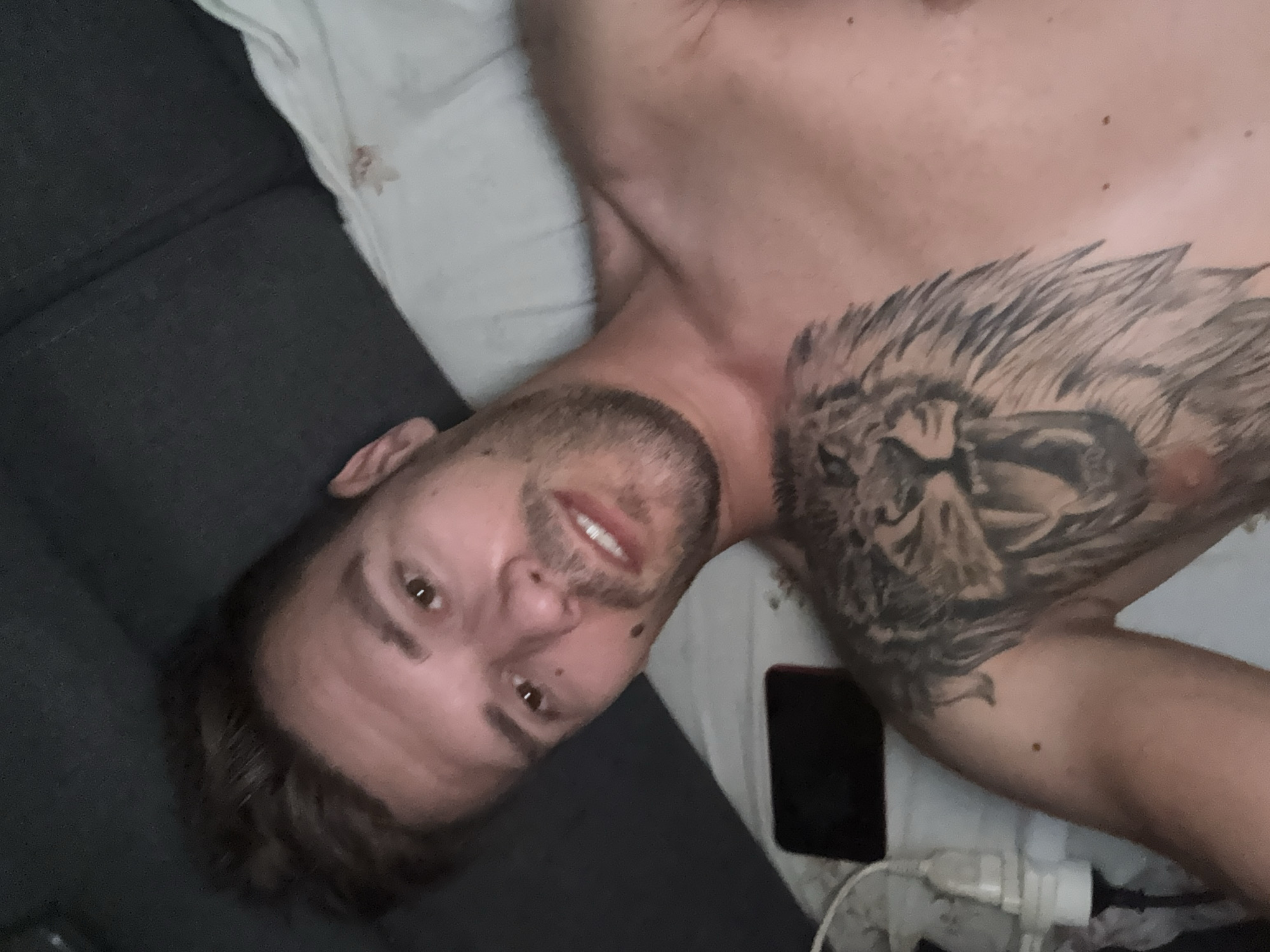The image is a close-up, high-angle selfie of a shirtless man lying on a bed. The man has short, brown hair, brown eyes, and stubble. He is smiling faintly. On his chest and left shoulder, he has a large tattoo of a lion's head. To the left of the man, a smartphone and a white electrical plug are visible on the bed. The background consists of a dark gray surface, likely a headboard or wall, and a white pillow. There are no discernible elements in the image that would indicate its location in Sesto Calende, Italy.