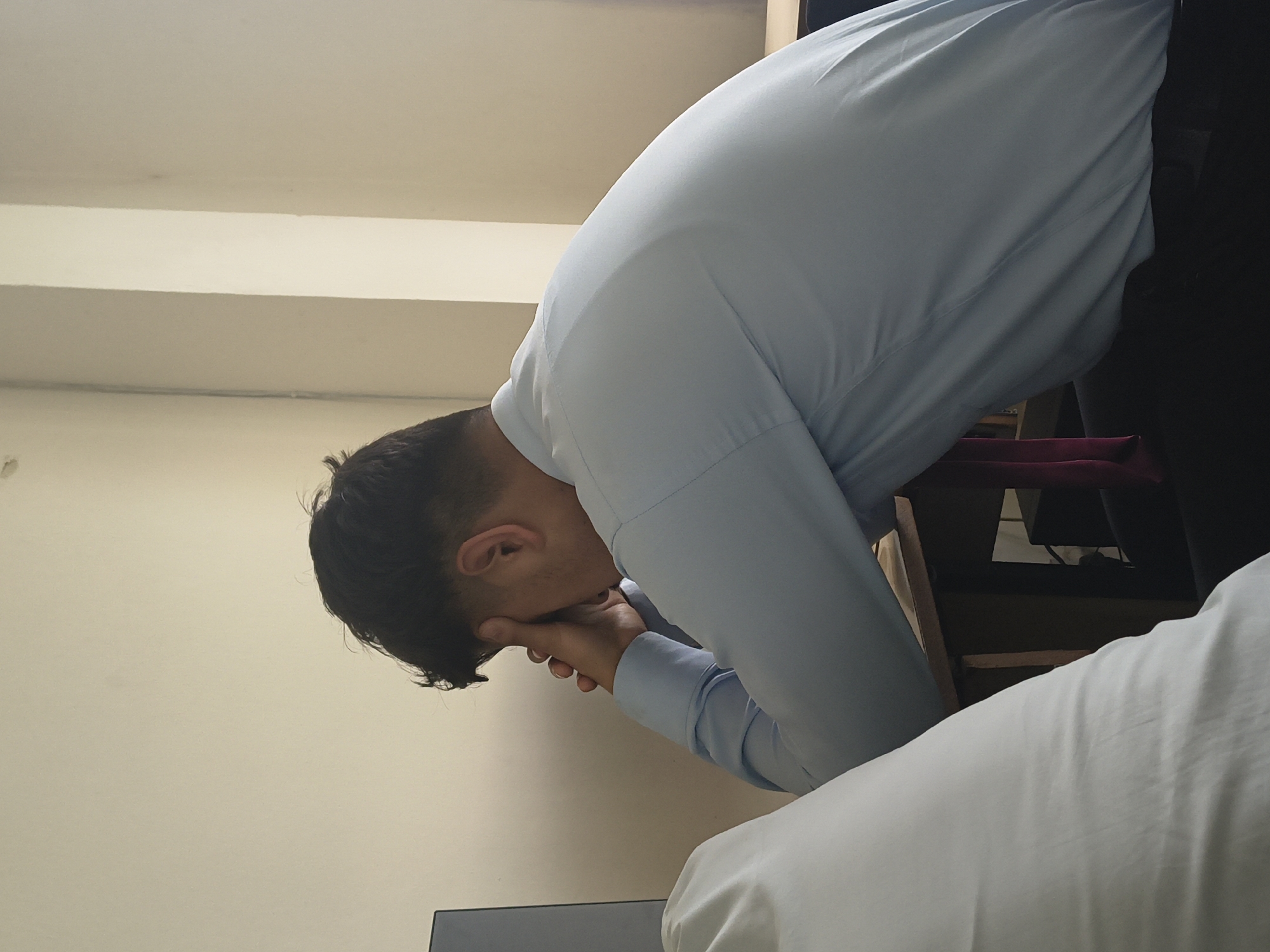 A young man in a light blue collared shirt is sitting at a desk with his head in his hands. His eyes are closed and he appears to be distressed or exhausted. The setting appears to be an indoor space, possibly a classroom or office, with neutral-colored walls and natural light. A white cloth, possibly a blanket or sheet, is visible in the foreground. A dark-colored object, possibly a computer monitor, is partially visible to the left. The man's hair is dark and styled with a fade on the sides. The overall mood of the image is one of quiet contemplation or struggle.