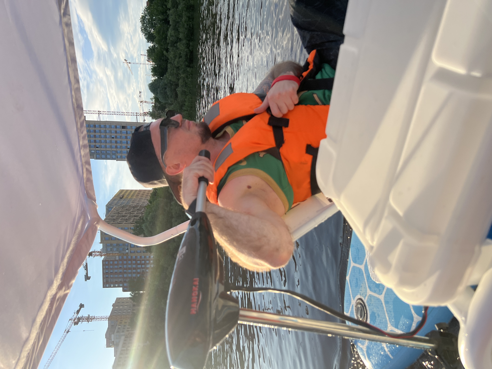 A man wearing sunglasses and a black baseball cap is seated on a boat, facing right. He has a life jacket on and is holding onto a trolling motor handle. In the background, there are several unfinished buildings under construction, as well as trees and a body of water. The sun is shining, casting a warm glow on the scene. The location is the Tura River in Tyumen, Russia, where there is ongoing urban development.
