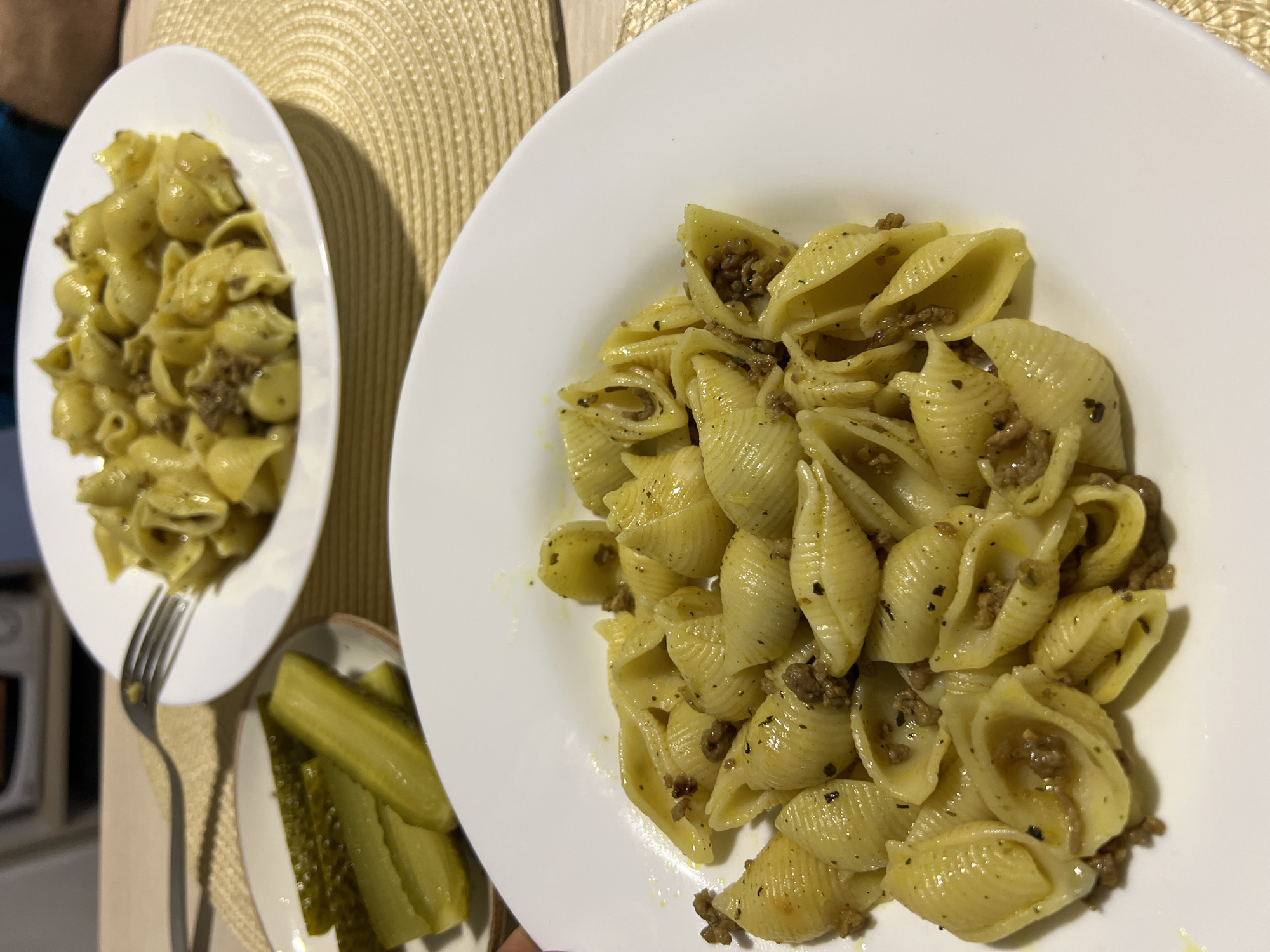The image presents two plates of pasta shells filled with meat, seemingly ground beef, on a woven placemat. A small dish of pickle spears sits next to one plate, accompanied by a fork. The setting appears to be indoors, possibly in a residential kitchen, with a stove visible in the background. There is no evidence to suggest this is in Boreti, Montenegro. The food is the primary subject, showcasing a meal prepared and presented for consumption.