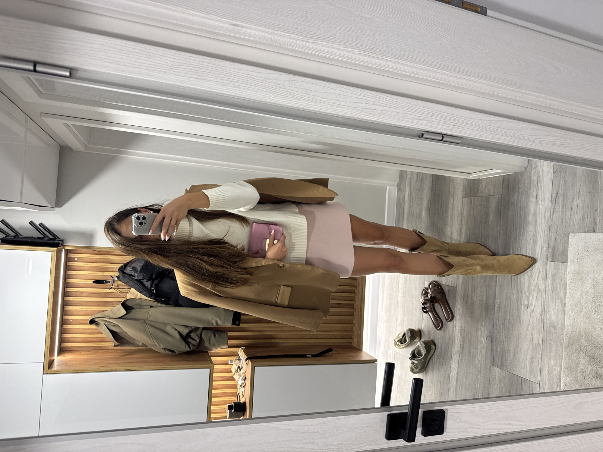The image depicts an indoor scene, likely a hallway or dressing area, with a woman taking a mirror selfie. She is wearing a white turtleneck sweater, a tan blazer draped over her shoulders, a pale pink mini skirt, and tan cowboy boots. She is holding a small pink clutch bag and a mobile phone.
In the background, there is a wooden panel with hanging coats, and a white cabinet with items on its surface. The floor is covered with grey wood-effect tiles. To the left, a black door handle is visible on a grey door. Several pairs of shoes are placed on the floor, including sneakers and brown slides. A white rug is partially visible at the bottom.
The image does not contain any information that would allow for identification of the location as Burgas, Bulgaria.