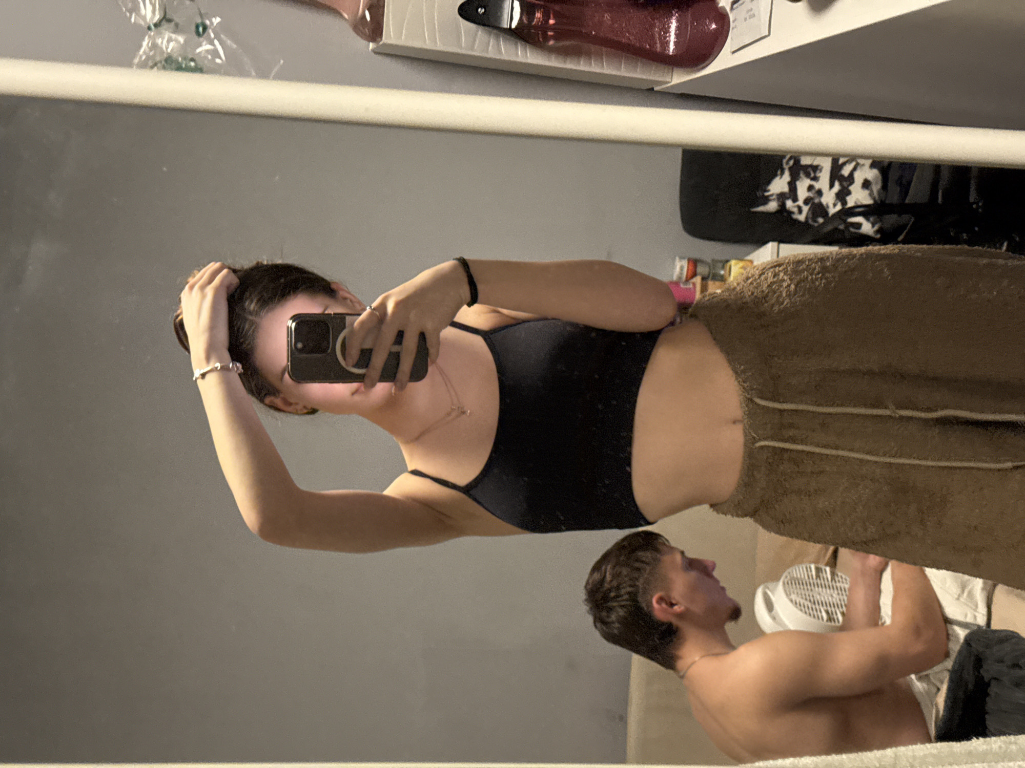 A woman in a black top and brown pants is taking a selfie in a mirror. The reflection shows a man shirtless lying on a bed, holding a white object. The location is impossible to determine from the given information.