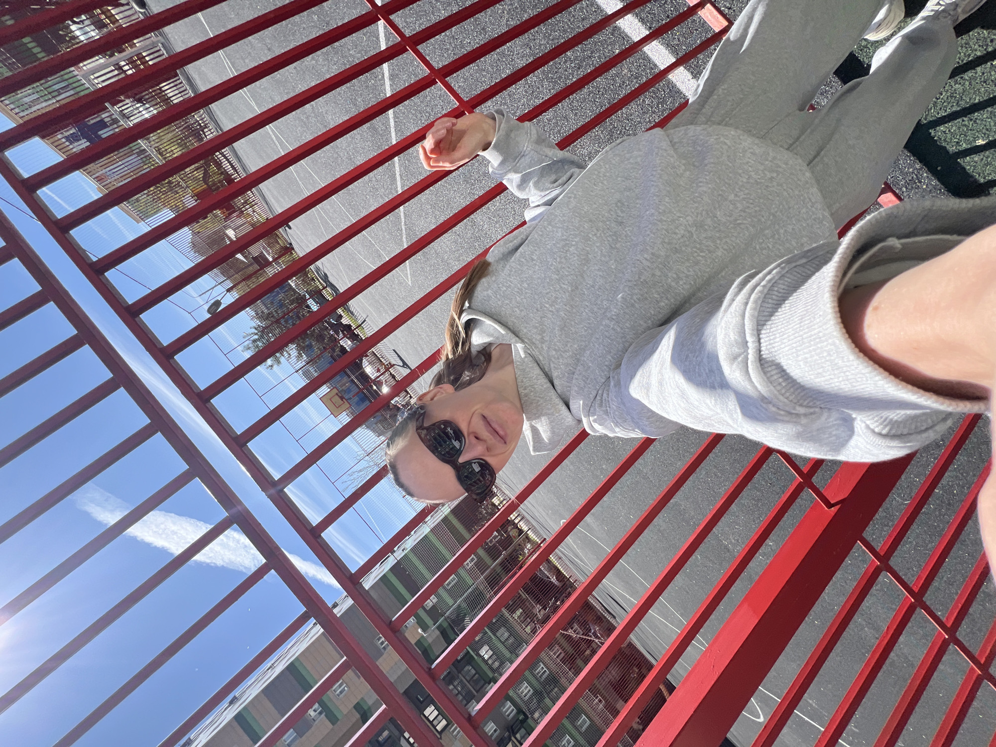 The image is a selfie of a woman taken outdoors on a bright, sunny day in Khimki, Russia.
**Subjects:** The primary subject is a woman, captured from a slightly low angle, looking towards the camera with a faint smile. She is wearing dark sunglasses.
**Objects:** She is dressed in a casual, matching grey hooded sweatshirt and grey sweatpants, paired with light-colored sneakers. The scene is dominated by a prominent red metal fence or set of bars, which spans the foreground and mid-ground, creating strong diagonal and horizontal lines across the frame. In the background, part of a basketball hoop and net are visible.
**Scene/Setting:** The setting appears to be an urban outdoor sports court or playground area. Behind the red fence, a grey paved surface, possibly asphalt, with faint white lines indicative of court markings, is visible. Beyond this, under a clear blue sky adorned with a few wispy white clouds, stand several multi-story buildings, likely residential or commercial, with diverse window patterns and some sections featuring green accents.
**Activity or event taking place:** The woman is taking a selfie, holding the camera herself, likely leaning against or standing near the red fence.
**Notable details:** The time of day is bright daylight, evidenced by a strong sun flare visible in the upper left portion of the image and clear shadows cast by the red bars onto the paved ground. The weather is sunny with a clear sky. The woman's expression is relaxed and casual. No visible text is present in the image.