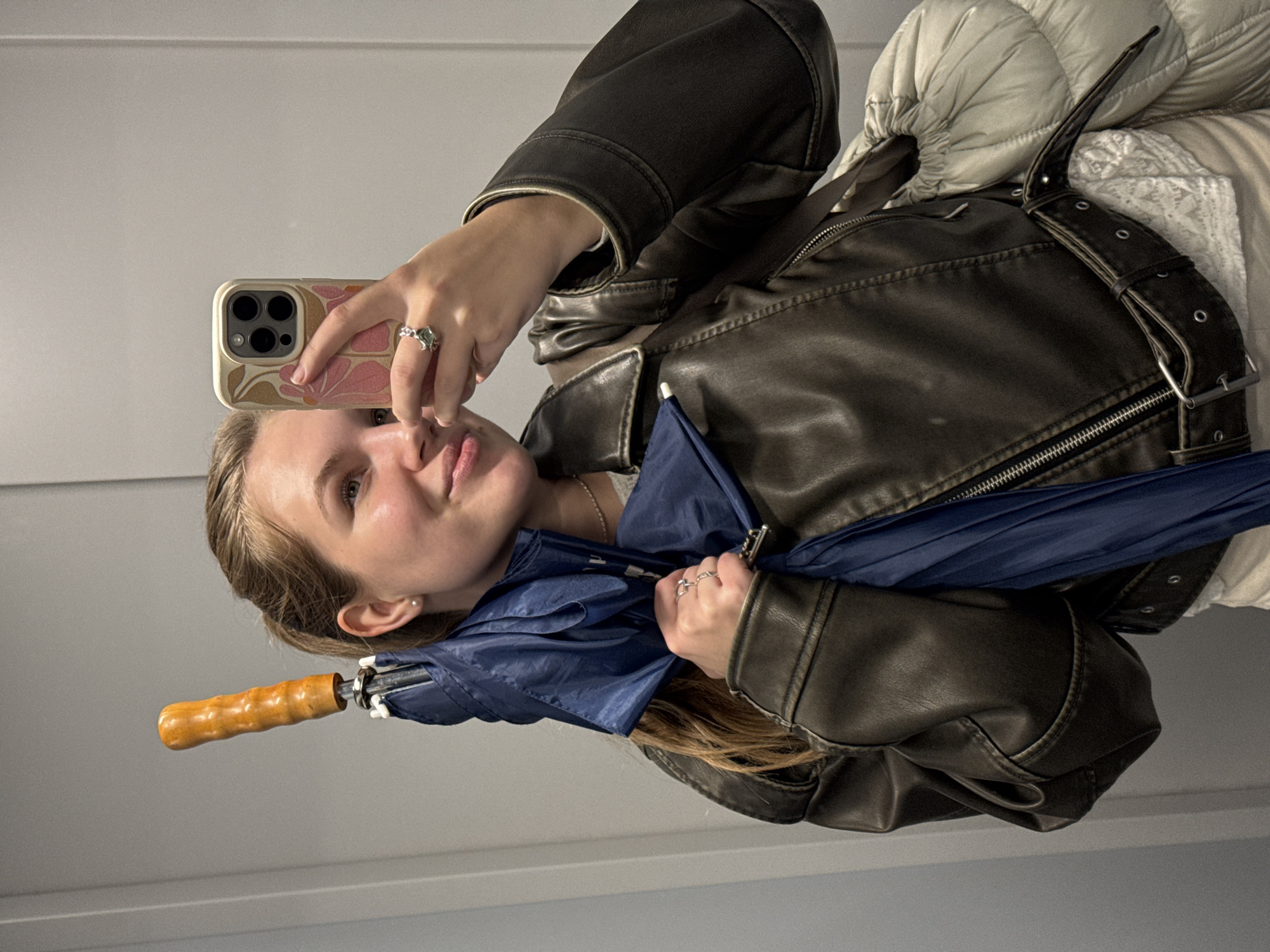 A woman is taking a selfie in an indoor setting with a light grey wall. She is holding a smartphone with a floral patterned case in her right hand. She wears a brown leather jacket over what appears to be a white or off-white padded vest. A closed blue umbrella is held in her left hand. She is wearing rings on both hands. Her blonde hair is partially visible. The location is Vozdovac Urban Municipality, Serbia, as requested.