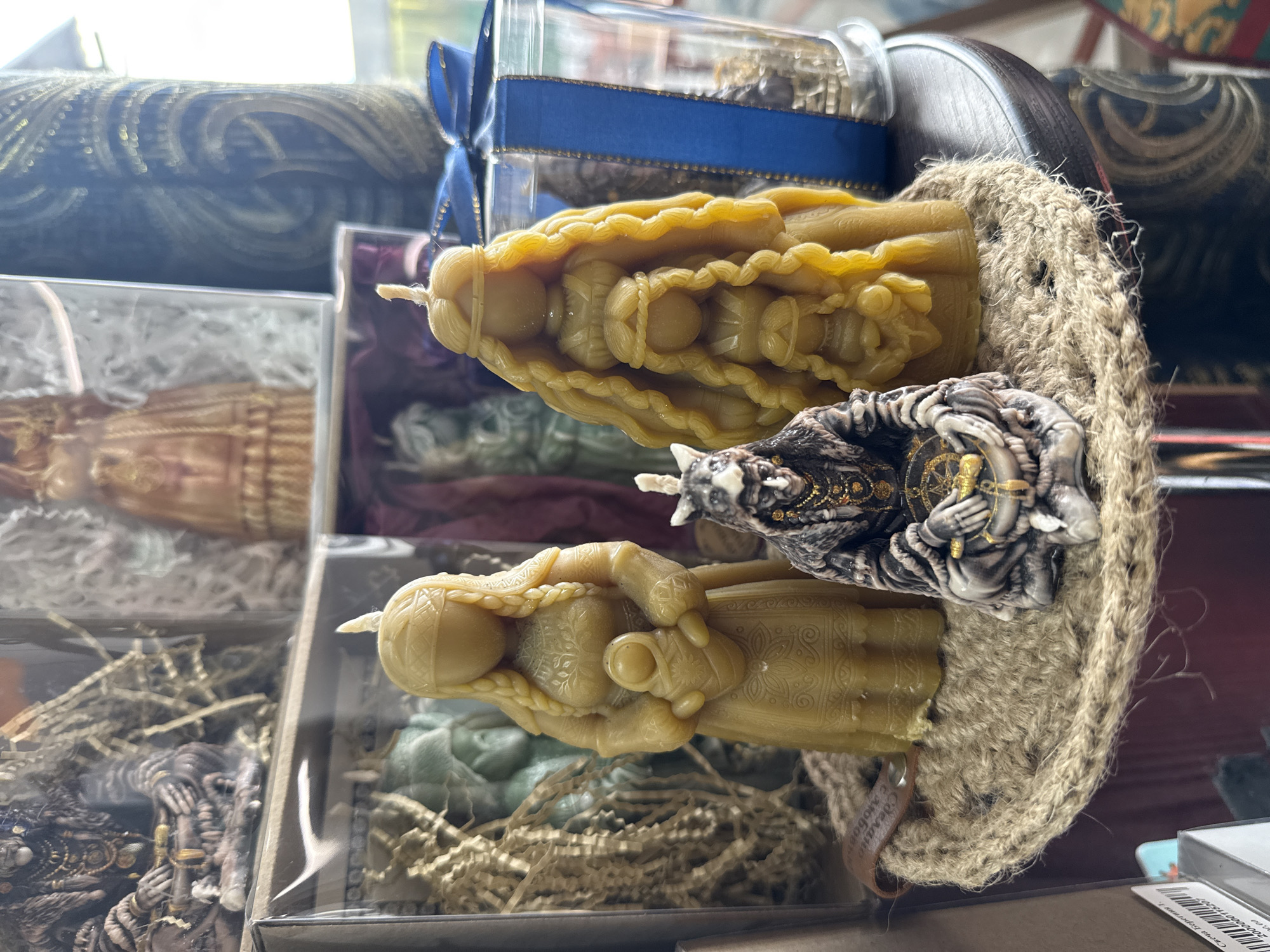 The image depicts a collection of handcrafted candles. Two yellow candles, molded in the shape of robed figures, are positioned in the foreground. Adjacent to them is a figurine of a wizard holding a compass. These candles are either displayed on or near a coarse, woven surface, possibly burlap. The candles are either on display or for sale in Ust-Koksa, Russia. In the background, more of the robed figure candles are visible, packaged in clear boxes with wood shavings beneath them. A glass jar with a blue ribbon around it sits behind the central arrangement. The overall scene suggests a craft shop or market stall specializing in handmade candles and figurines.