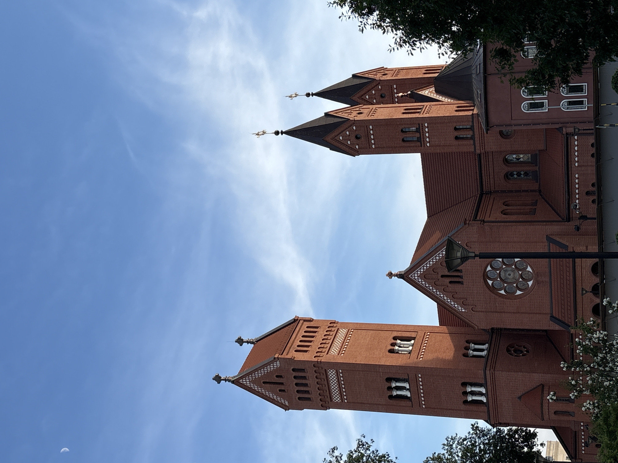 The image shows the Church of Saints Simon and Helena, also known as the Red Church, located in Minsk, Belarus. The church is made of red brick and features a tall tower with a steeple and several windows. The roof is red tile. The sky is blue with some wispy white clouds. A crescent moon is visible in the upper left corner of the sky. The church is surrounded by trees and foliage. A lamppost is positioned in front of the church. The overall scene is a clear day with good visibility.
