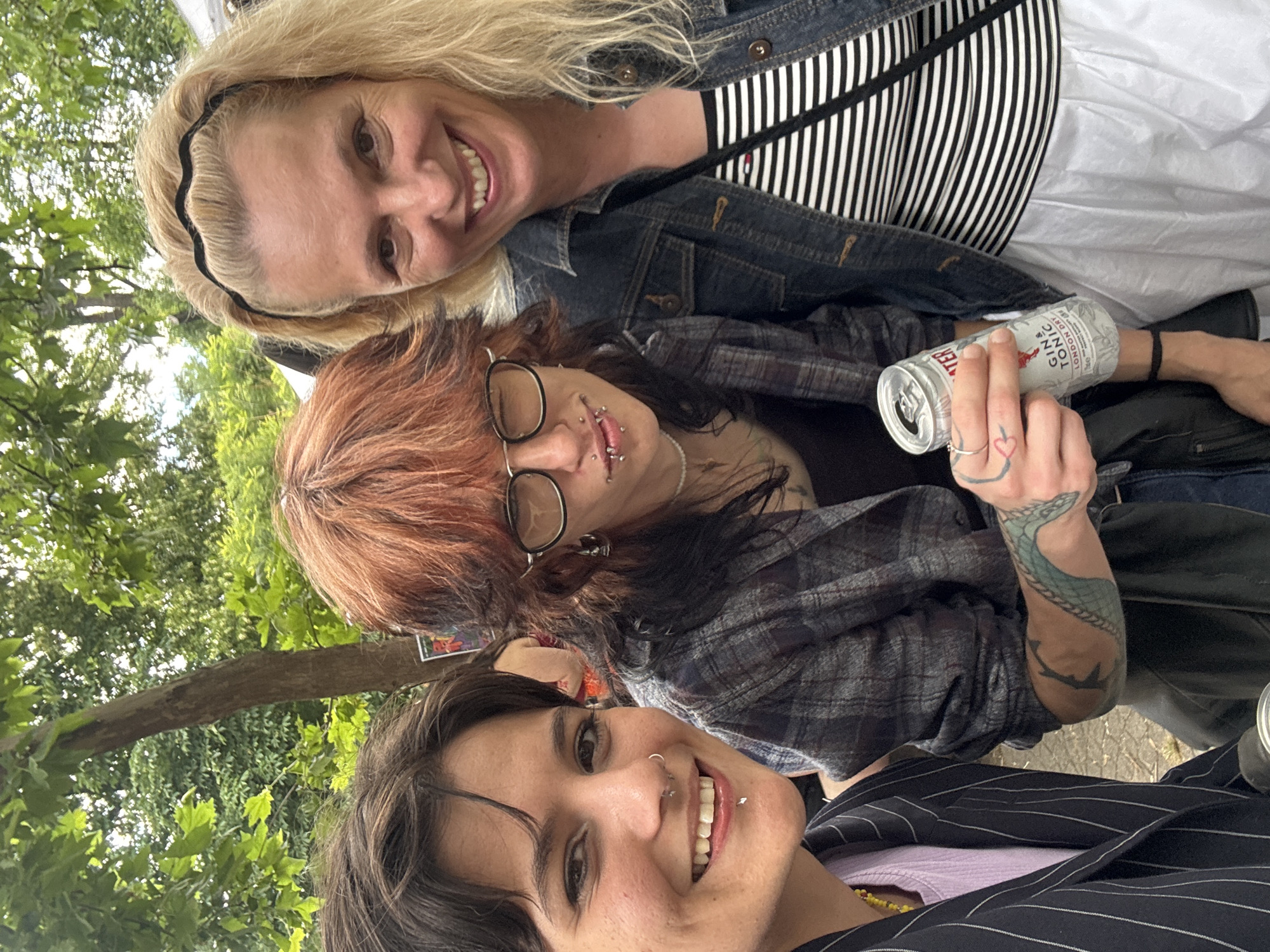 Three individuals are posed for a selfie outdoors, with trees and foliage in the background, suggesting a park or garden setting. The person on the left, with short dark hair and a nose ring, is smiling and wearing a pinstriped jacket over a lavender top. The person in the middle, with reddish-brown hair, glasses, and facial piercings, is holding a can of "GIN & TONIC LONDON DRY" and wearing a plaid shirt. The person on the right, with blonde hair and a black headband, is smiling and wearing a denim jacket over a black and white striped top. The lighting indicates it is daytime. No specific city landmarks of Sofia, Bulgaria are identifiable in the image.