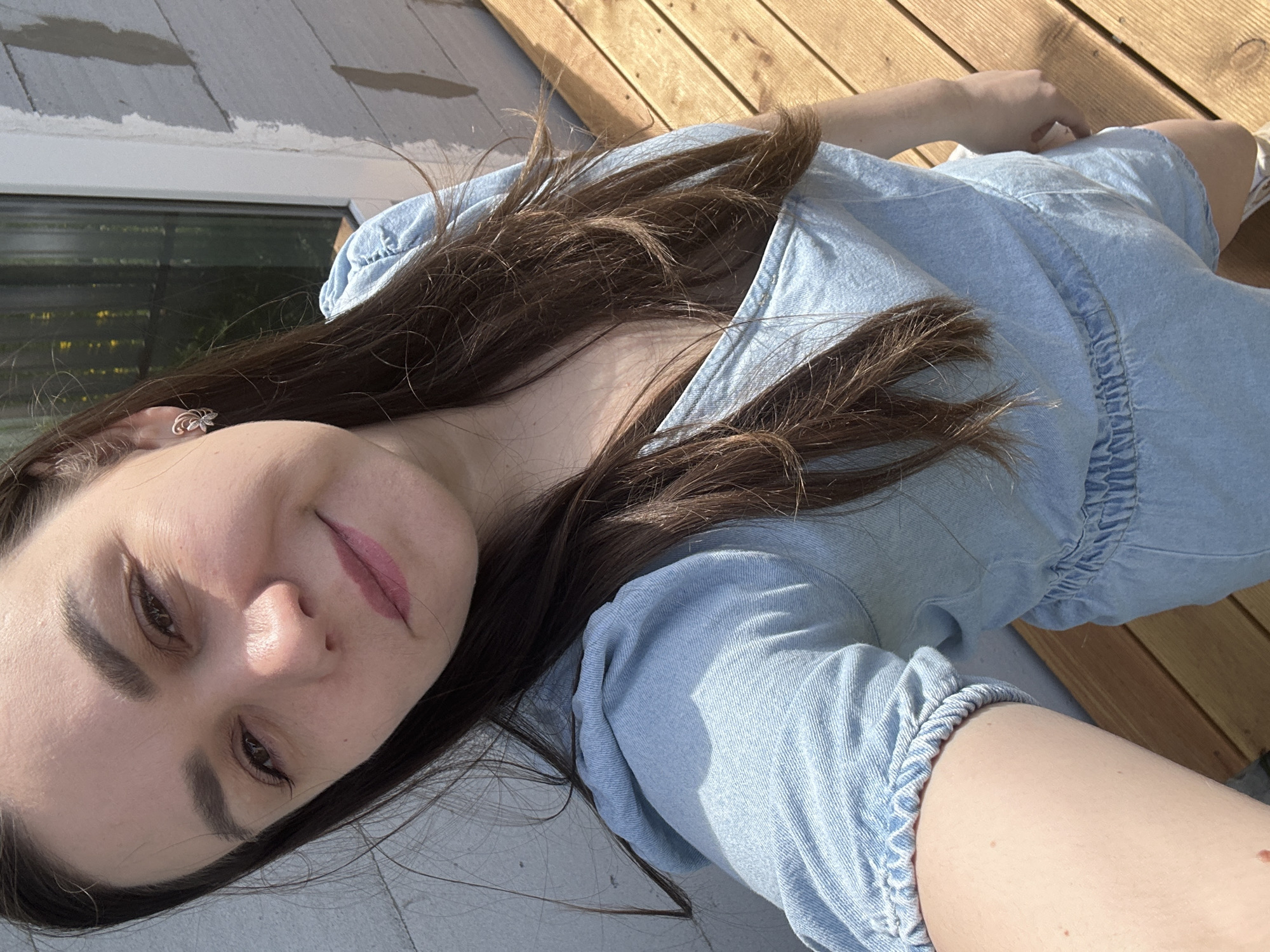 A woman with long brown hair is taking a selfie while lying on a wooden deck. She is wearing a light blue denim dress. A building with windows is visible in the background. The location is possibly in the city of Chaikovsky, Russia.