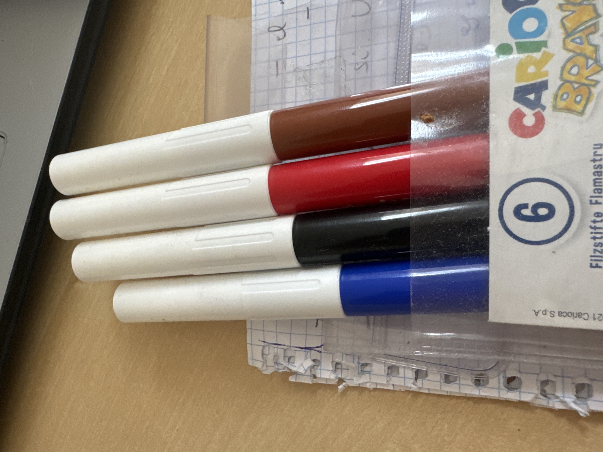 The image shows a close-up, angled view of four markers in a clear plastic case, resting on a wooden surface. From left to right, the markers are blue, black, red, and brown. A torn piece of graph paper with some writing and blue ink marks is partially visible beneath the markers. The background shows a blurred view of what appears to be a laptop or similar electronic device. There is no indication of the city of Murcia, Spain, in the image.