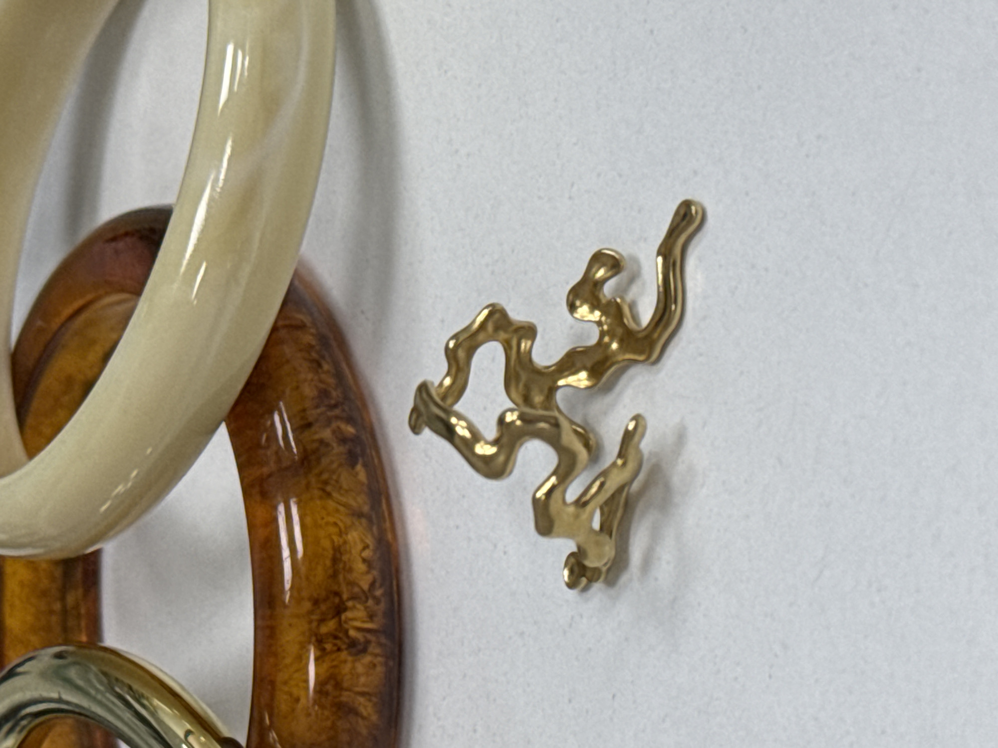 The image shows a close-up of several chunky, abstract-shaped jewelry pieces. On the left, there are two large, smooth, curved rings. The uppermost ring is a pale, creamy beige color, and the one beneath it is a translucent, rich amber or tortoiseshell pattern. They appear to be made of resin or a similar material. Partially visible at the bottom is another ring, this one appearing to be metallic, possibly gold or brass, with a similar chunky, curved design.
To the right of the rings, a small, intricately shaped gold charm or pendant is visible. It has an organic, almost dendritic or coral-like form, with multiple twisting branches and loops. The charm is also metallic and appears to be polished, reflecting light.
The background is a plain, light-colored surface, likely a countertop or table, with a subtle speckled texture. The lighting suggests an indoor setting, with the light source creating highlights on the glossy surfaces of the jewelry. No people, activities, or text are visible in the image. The focus is entirely on the details and textures of the jewelry pieces.
