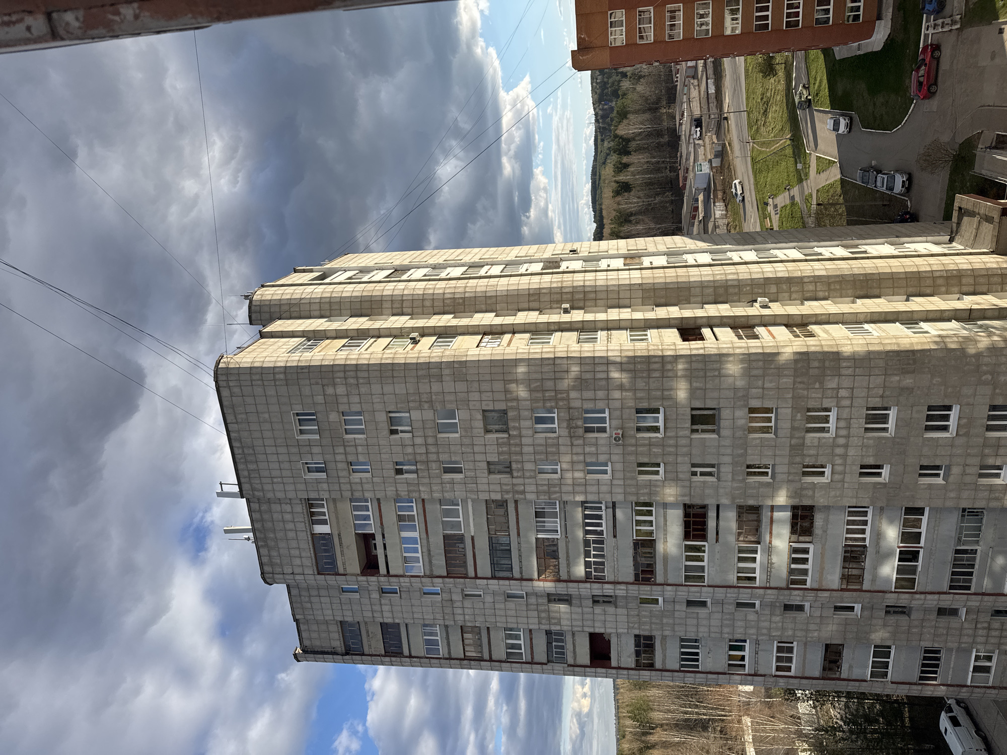 The image presents a high-angle view of an urban or suburban landscape in Chaikovsky, Russia, taken from a building overlooking the scene. A prominent, tall, multi-story concrete apartment building, likely residential, stands vertically in the mid-ground. Its facade is composed of light grey panels or tiles, featuring numerous windows and enclosed balconies. Some windows have a uniform design, while others show varied enclosures and minor signs of wear or modification, with several air conditioning units attached to the exterior. Antennas or communication equipment are visible on the building's roof.
To the right, a section of a reddish-brown brick building is visible, also with multiple windows. The background reveals a vast expanse of forest or treeline stretching across the horizon, with roads and grassy areas discernible between the structures. The sky overhead displays a dramatic contrast of heavy, dark grey clouds and patches of bright blue sky, indicating changeable weather. Power lines crisscross the upper portion of the frame.
At ground level, paved roads and parking lots are visible, populated by several parked cars, including a red vehicle and multiple white ones. A small yellow excavator is present on a grassy patch near the road, suggesting ongoing work or daily activity. The lighting indicates it is daytime, with distinct shadows. No people are clearly visible, and no discernible text is present in the image.