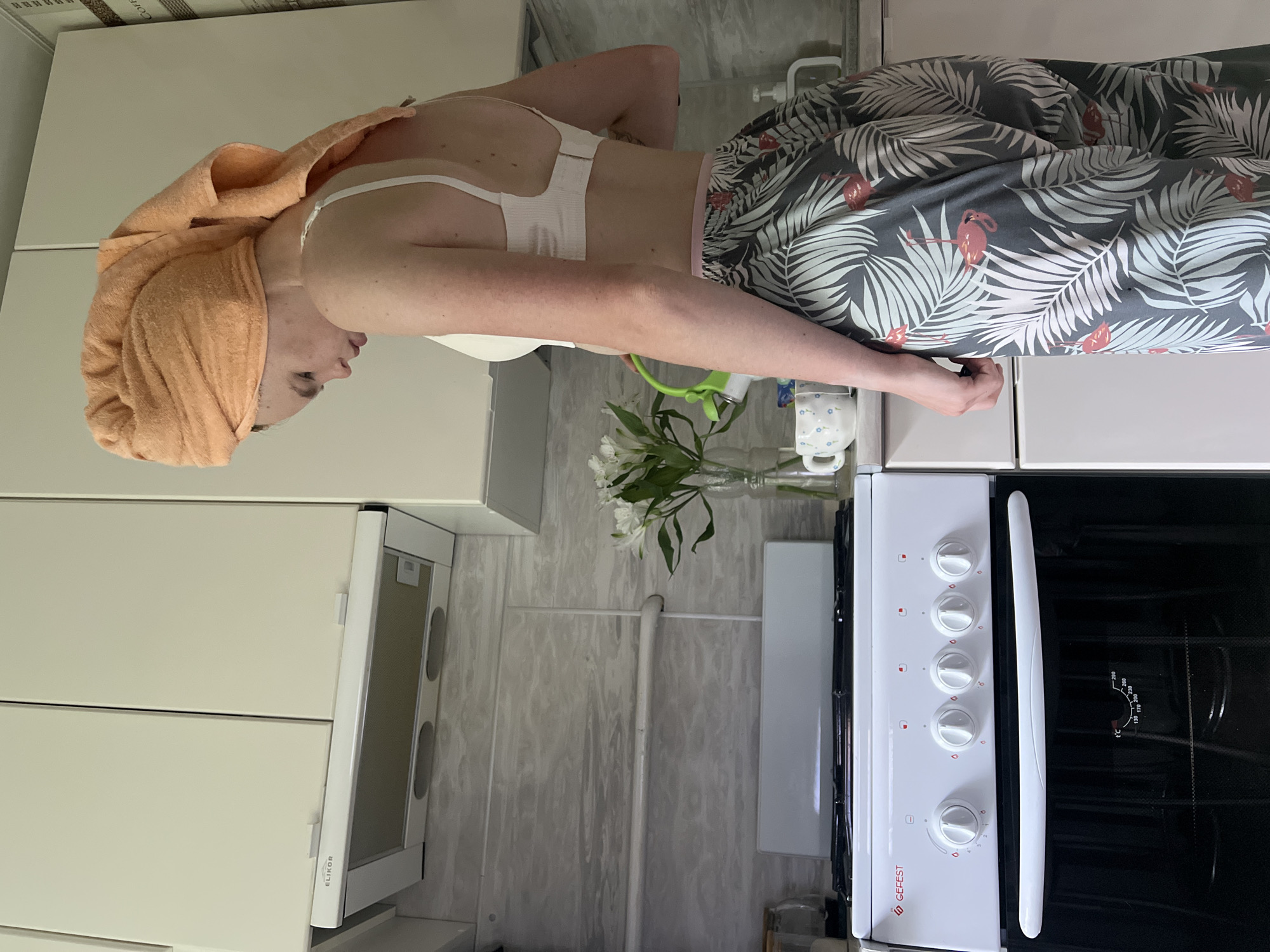 In a domestic kitchen, likely located in the city of Chaikovsky, Russia, a young woman with an orange towel wrapped around her head and wearing a white bra and patterned pajama pants stands by a stove. She is in the process of watering a vase of white flowers. The kitchen features white cabinets above a light-colored backsplash. A white gas stove with five knobs and an oven is visible on the left. On the counter, to the right of the flowers, sits a white mug with a floral pattern. The overall scene suggests a mundane, everyday activity in a home environment.