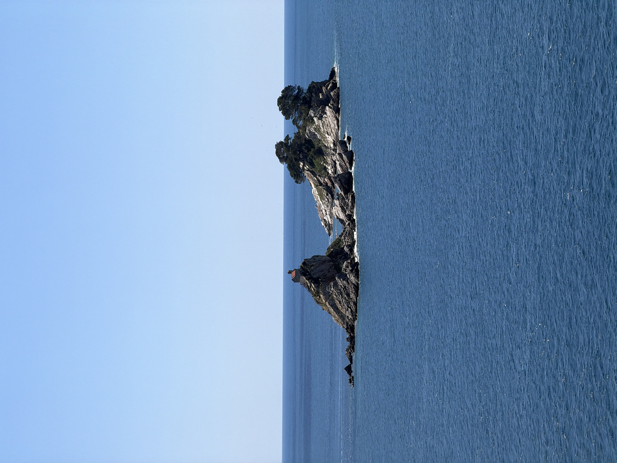 The image shows a view of the Kastio Peninsula in Petrovac na Moru, Montenegro. The peninsula is a rocky outcrop with trees and shrubs growing on it. A small, orange-roofed structure is located on the higher part of the peninsula. The surrounding environment consists of blue sea on the right and a light blue sky and horizon on the left. The water surface has a subtle texture.