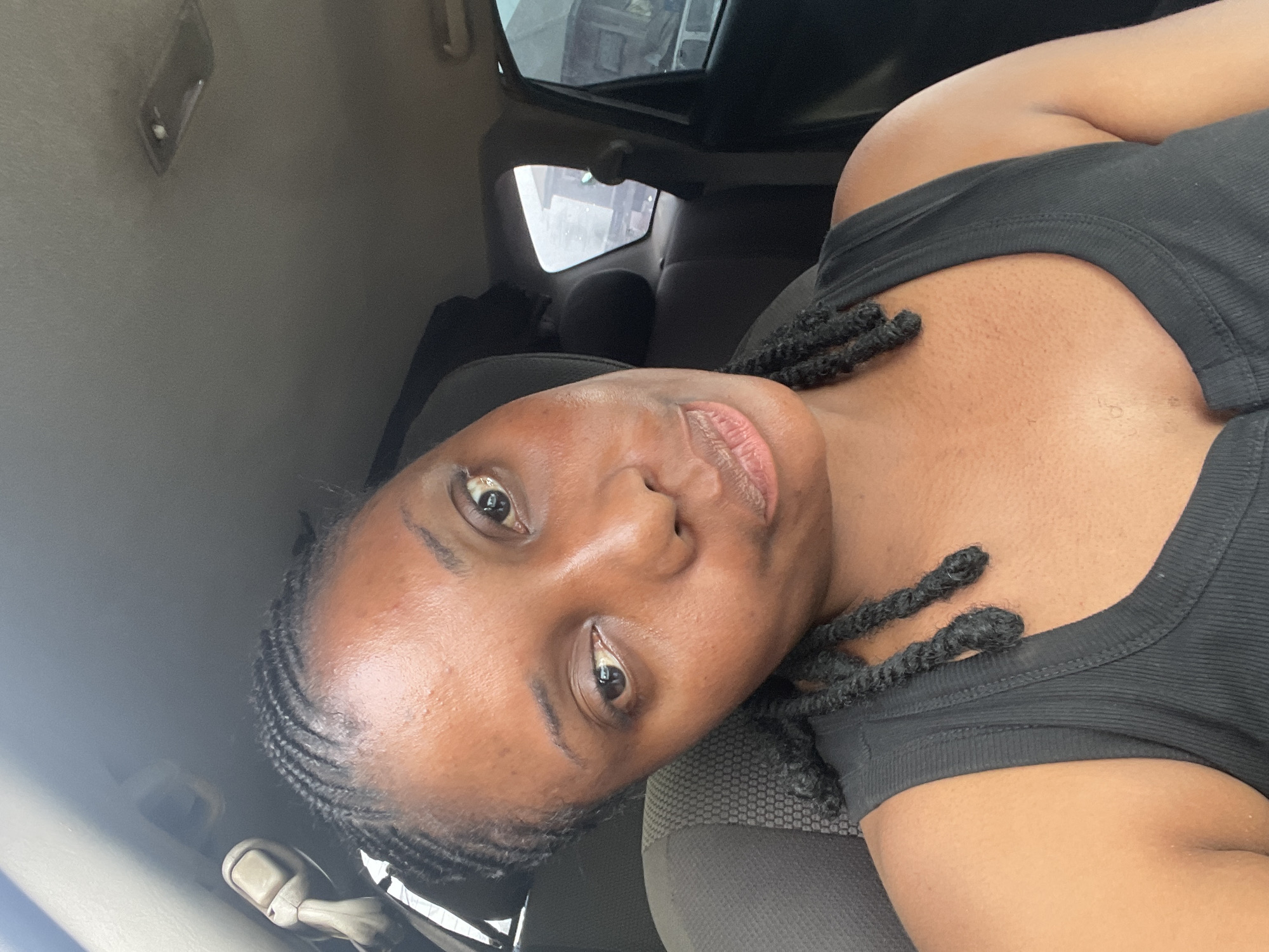 A woman with dark skin and braided hair is sitting in the passenger seat of a car. She is wearing a black tank top. The interior of the car is gray. The view out the window is blurred, but appears to be a street scene. There is no information to confirm the location as Jos, Nigeria.