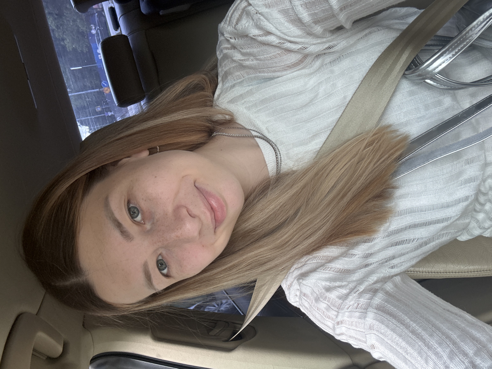 A woman is in a car, wearing a white shirt and a seatbelt. She has light brown hair and freckles. The car appears to be moving, with a view of a street and buildings visible through the window. There is no clear information to indicate the specific location of the city of Moscow, Russia.
