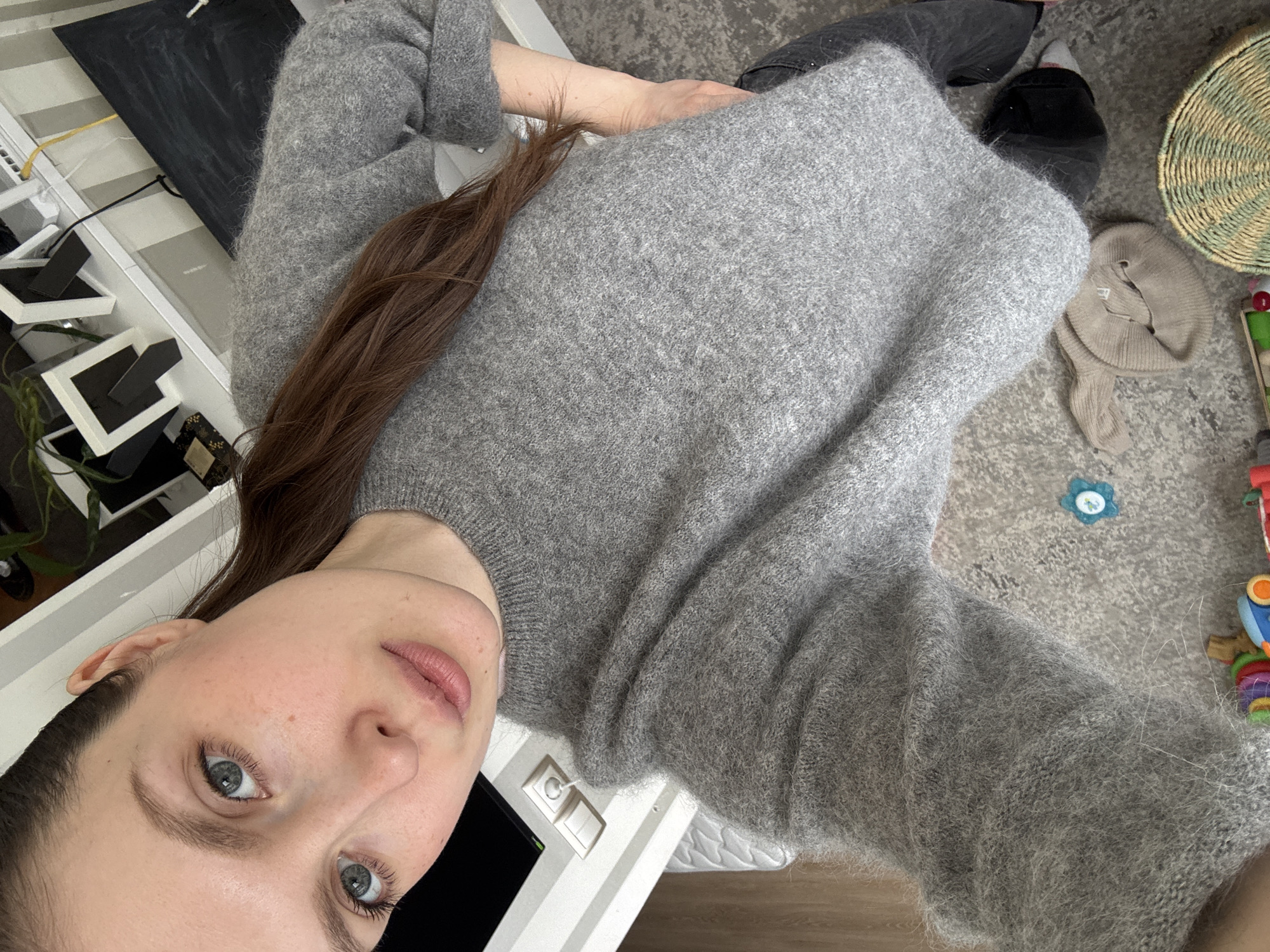 A woman with long brown hair and blue eyes is seen from a high angle. She is wearing a gray sweater and looking directly at the camera. The background includes a white shelving unit with various items, including framed pictures and plants. The floor is visible, with a wicker basket, a beige hat, and children's toys scattered around. The scene appears to be indoors, possibly a residential setting. The location is not identifiable as being in Moscow, Russia.