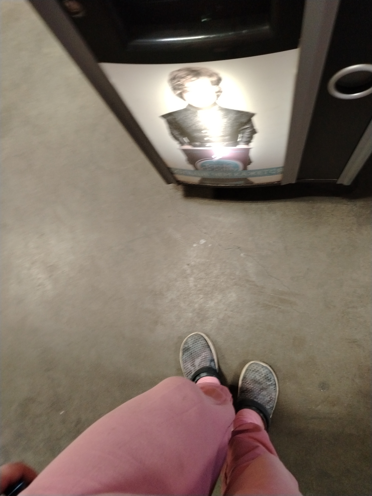 A high-angle, first-person shot captures a person's lower body standing on a plain, light grey floor, facing an upright display or machine. The person is wearing pink trousers and dark grey, textured slip-on shoes. In the background, a screen or reflective surface within a dark machine displays an image of a human figure. This figure, likely male, has light brown hair and is depicted from the chest up in dark, possibly armored attire. The figure's face and upper body are intensely illuminated, causing overexposure that obscures specific features. In front of the figure, a blue, curved object with faint, light-colored text, possibly in Cyrillic script, is visible but largely illegible due to blurriness and glare. The setting appears to be an indoor public space, such as a store or an arcade. No specific activity is depicted beyond standing, and the time of day and emotions are not discernible.