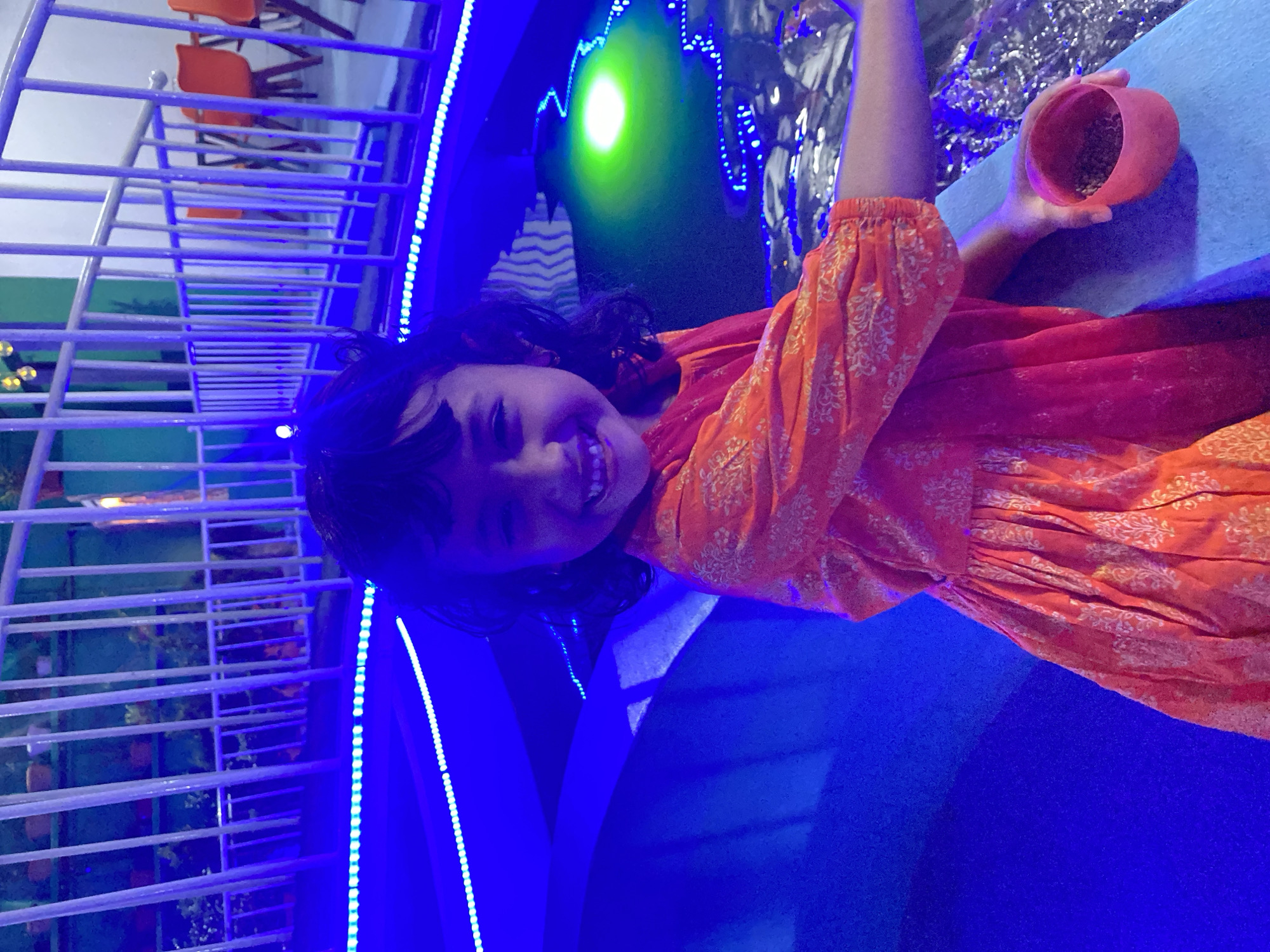 The image depicts a young girl in Dhaka, Bangladesh, smiling while holding a small, terracotta pot. She is wearing an orange dress with a white floral pattern. The background includes a railing with blue LED lights and a body of water with light reflections. There are also chairs visible in the background. The scene appears to be indoors, possibly at a park or recreational area in Dhaka.