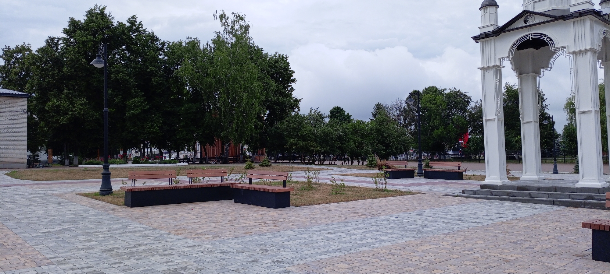 The image depicts a paved public square in the city of Kirsanov, Russia, featuring a white architectural structure with arched openings and columns, likely a gazebo or monument. Several park benches with wooden slats and dark bases are strategically placed throughout the square, some in L-shaped configurations. A lamppost with a decorative base and a single light fixture stands near the center-left of the frame. Lush green trees, including a tall birch tree, line the background, with a brick building visible on the far left. The sky is overcast with heavy clouds. The ground is paved with interlocking stones in varying shades of grey and tan, with patches of dry grass and small shrubs surrounding the benches. The scene appears to be unoccupied by people.