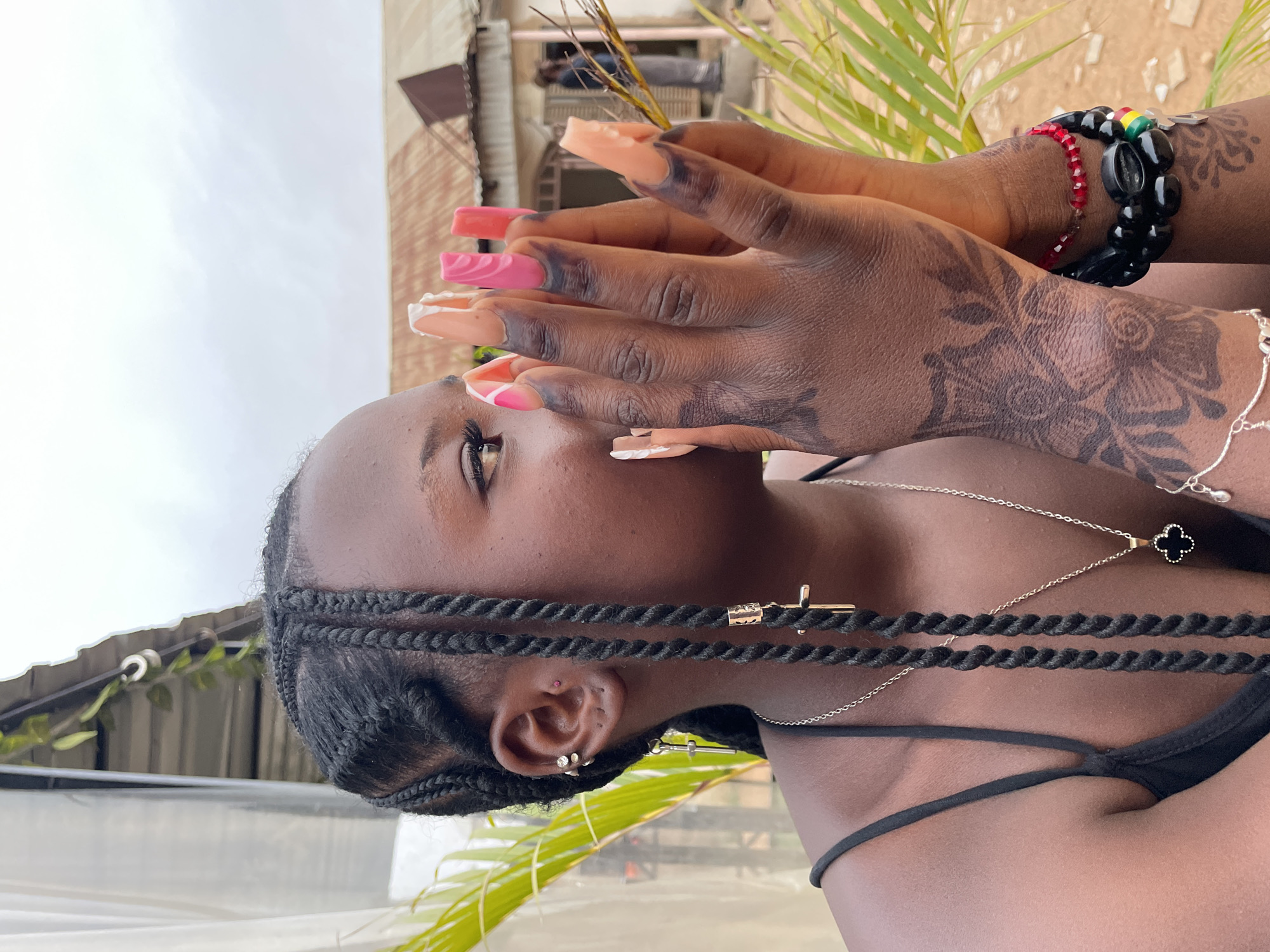 A young Black woman in Jos, Nigeria, is photographed from a low angle. She wears a black tank top and her hair is styled in braids adorned with gold-colored accents. She wears multiple necklaces and earrings, and her hands are raised to cover part of her face. Her hands are decorated with henna tattoos. She has long, artificial nails with a variety of pink and white designs. One arm has a bracelet made of beads. The background includes vegetation, buildings, and a gray sky.