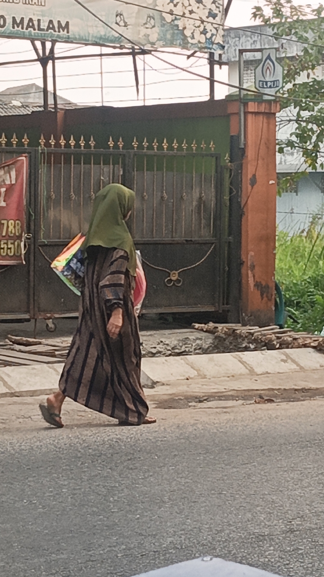 A person wearing a green hijab and a striped brown and black dress walks along a sidewalk in Banjarmasin, Indonesia. The person is carrying two shopping bags, one in their right hand with a colorful geometric pattern and another pink bag in their left arm. The person is wearing flip-flops. In the background, there is a metal gate with ornate decorative elements, a sign that says "ELPIJI" with a logo, and a building with orange and green paint. Vegetation is visible behind the building. The ground appears to be asphalt and concrete.
