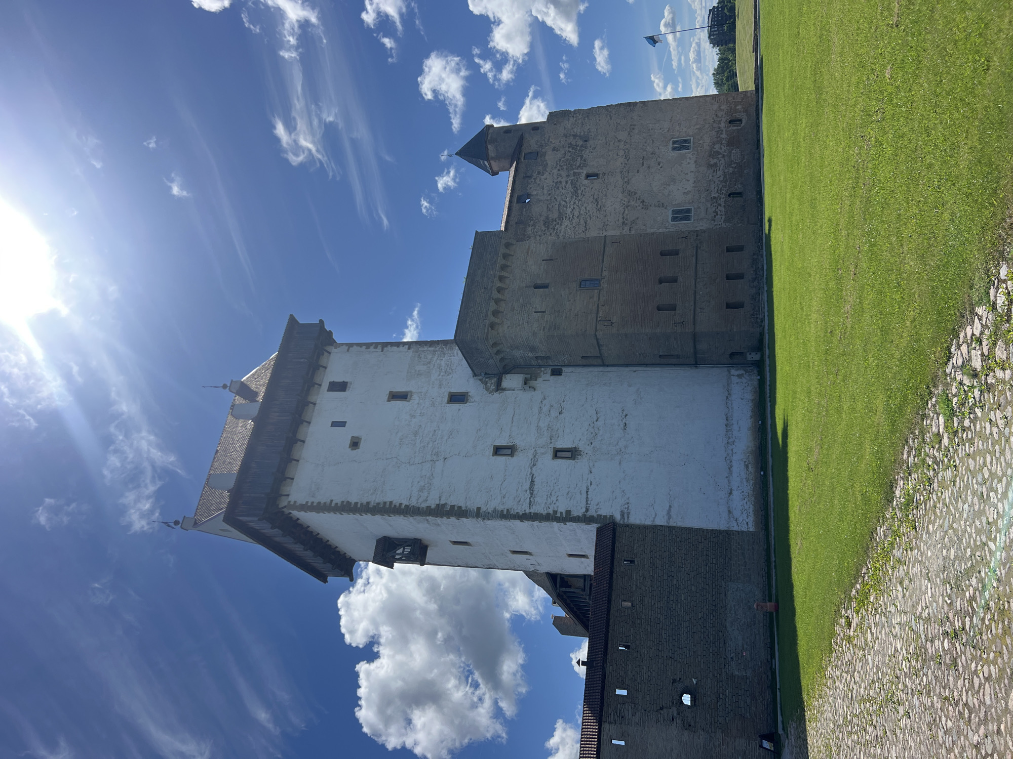This is a vertical, eye-level, outdoor, daytime shot of the Narva Castle in Narva, Estonia. The castle is a large, stone structure with a white-plastered tower and a darker, brick-like base. It features several windows of various sizes and a dark, pitched roof on the tower. To the right of the castle is a well-maintained, bright green lawn. In the foreground, a cobblestone path leads alongside the grass. The sky is a vibrant blue with scattered white clouds, some of which are wispy and streaky, indicating a bright and sunny day with a gentle breeze. The sun is visible in the upper left corner of the image, casting a bright glare and lens flare. A flag is visible in the distance flying from a pole on top of a hill behind the castle. The overall mood is peaceful and serene, showcasing a historical landmark on a beautiful day.