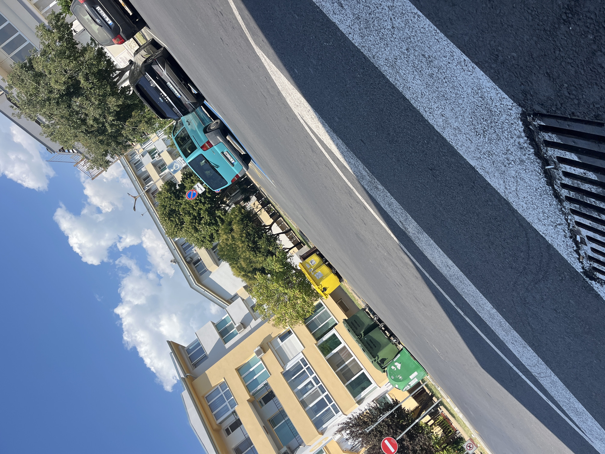 The image depicts a street scene in the city of Pomorie, Bulgaria, under a bright blue sky with scattered white clouds. In the foreground, the asphalt road features prominent white pedestrian crossing stripes and a street drain. To the left, a building with a yellow facade and white trim, adorned with numerous windows and balconies, lines the street. Adjacent to the building are several green and one yellow garbage bins. Further down the street, a turquoise minivan is parked, followed by a black SUV and another black car. Trees with lush green foliage are visible between the buildings and along the sidewalk. A red and white circular traffic sign is also present on the left side of the road. The image is captured from a tilted perspective.