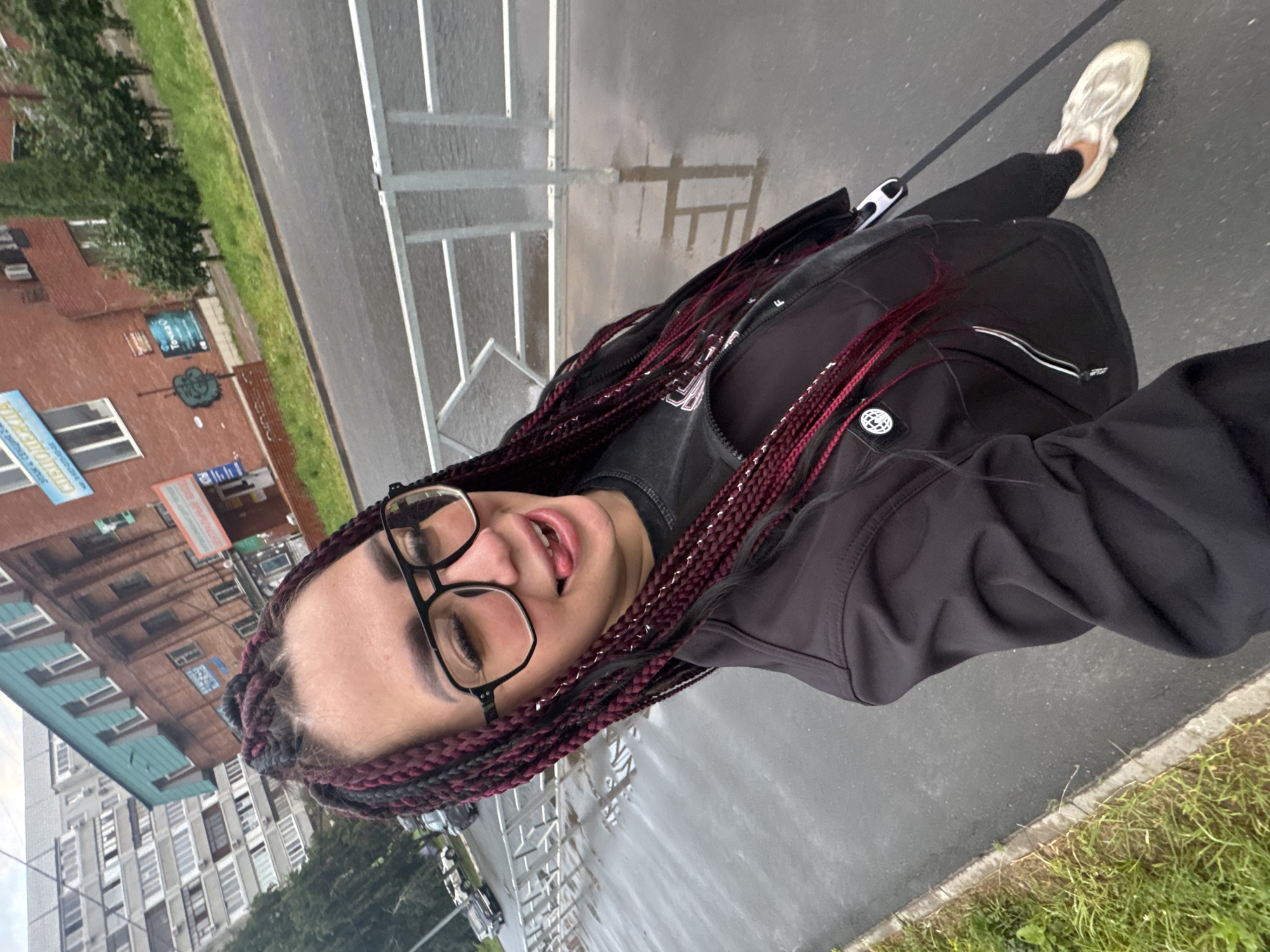 A woman with burgundy braids and glasses is taking a selfie outdoors on a wet, overcast day in Tolyatti, Russia. She is wearing a black jacket over a dark t-shirt and light-colored shoes. Her tongue is sticking out of her mouth. In the background, there are brick buildings, a modern apartment building, trees, a metal fence, and cars on a road. The ground is wet, indicating recent rain. The scene appears to be a street or sidewalk area in a residential or mixed-use part of the city.