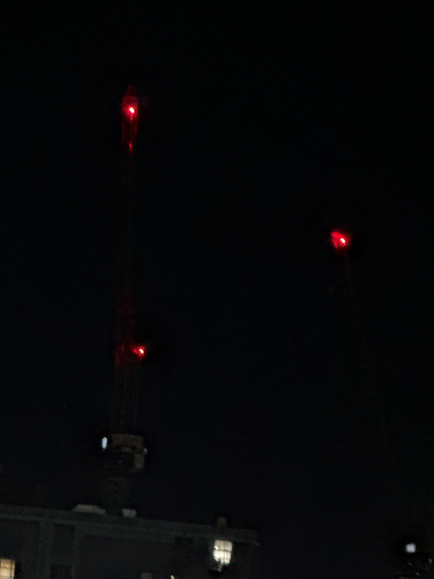 The image, taken at night in Basildon, United Kingdom, shows a dark urban landscape. In the foreground, the top of a building with illuminated windows is visible. Above the building, three red lights are prominent. Two of these lights appear to be part of a construction crane, with one positioned higher up and another on a lower section of the structure. The third red light is located further to the right and slightly higher than the crane lights, suggesting another elevated structure or beacon. The overall scene is characterized by deep shadows and the stark contrast of the red lights against the dark night sky.