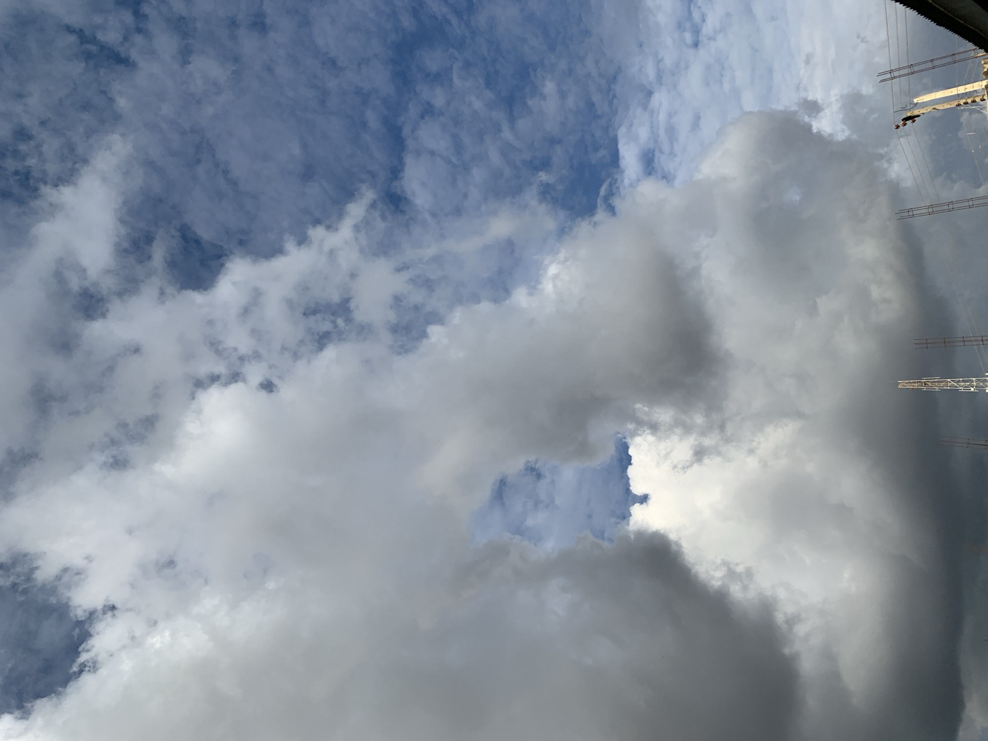 The image displays a dynamic sky over New Stadium, Nigeria, characterized by varying cloud formations. Dense, greyish-white cumulus clouds dominate the lower and central left frame, presenting an irregular opening that reveals a patch of lighter blue sky in the upper-middle left. The upper right quadrant exhibits a deeper blue sky with scattered, finer cloud patterns. In the lower portion of the image, several anthropogenic structures are visible. A white lattice telecommunications tower is partially visible in the lower-left-center. To the lower-right, multiple vertical rebar structures indicate ongoing building construction. Further right, a section of a dark-colored roof, a utility pole, and associated power lines are present. A single, small, dark human figure is discernible near the utility pole and roof, suggesting human activity related to these structures.