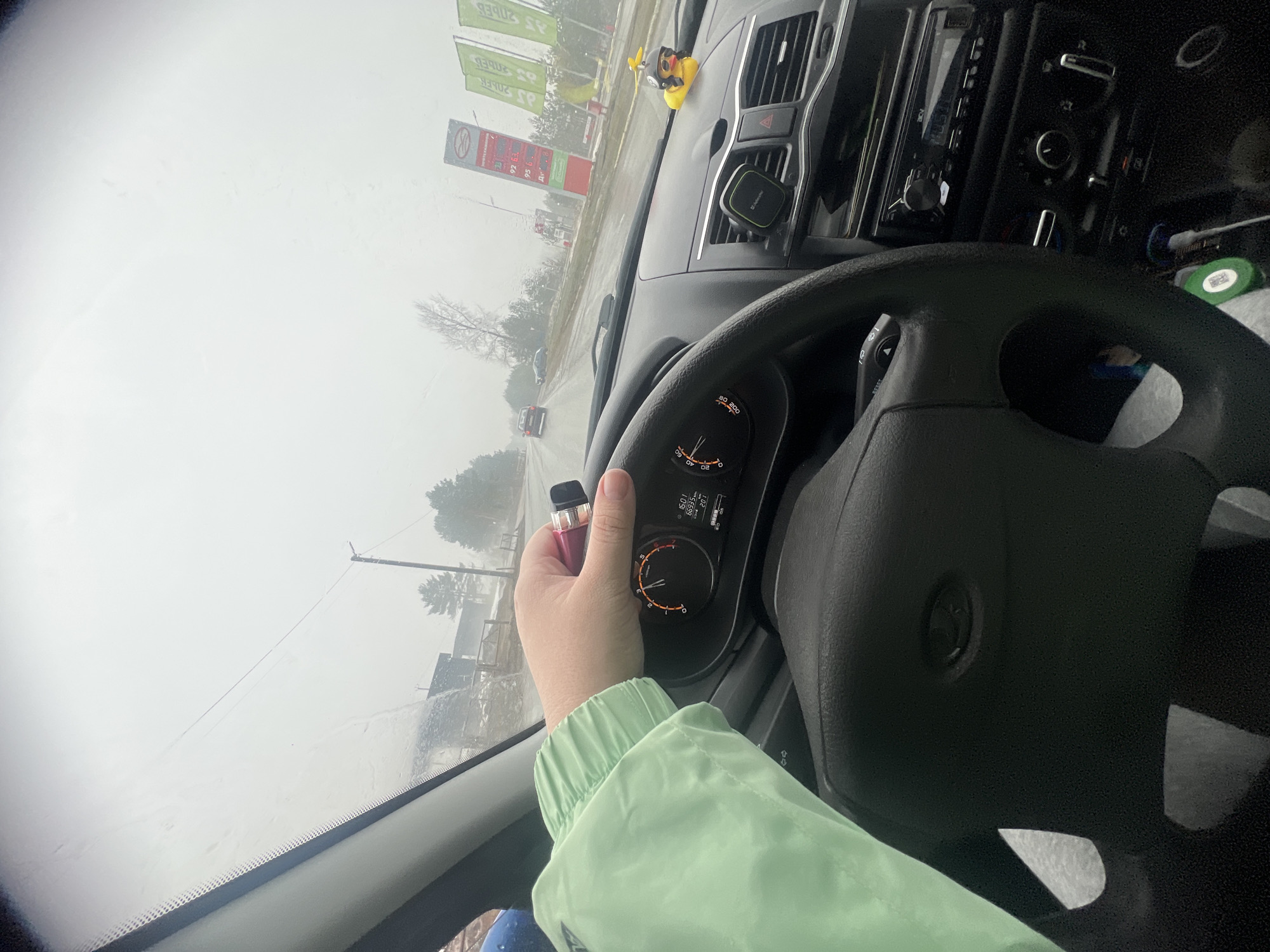 The image depicts the interior of a vehicle, likely a car, from the driver's perspective. A hand is holding a vape pen near the steering wheel, obscuring part of the dashboard, which displays information such as the time (16:01), mileage (66935), and temperature (40). Outside the windshield, the visible environment suggests a rainy or snowy day, with a gas station visible in the distance. The text "92" and "95" are visible on a red sign in front of the gas station, likely referring to fuel grades.
The gas station is identified as belonging to the "Lukoil" network based on the logo on the sign, a Russian petroleum company. The location is likely in the city of Chaikovsky, Russia, based on information from the OCR extracted text.