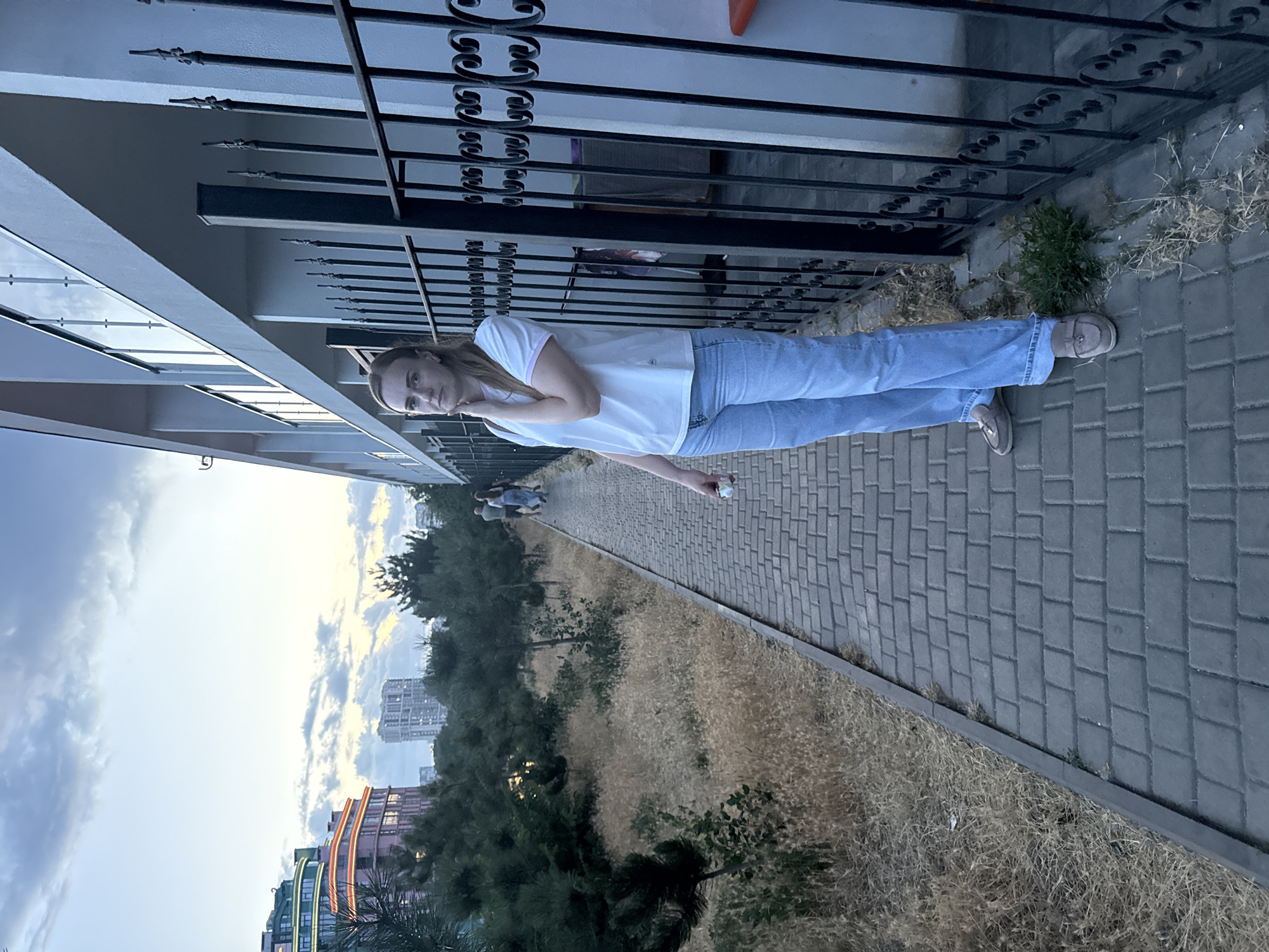 A full-body, slightly low-angle shot captures a woman standing on a paved walkway in Odesa, Ukraine, with a building and a black metal fence to her right, and dry, overgrown vegetation and trees to her left. The woman, facing the camera with her left hand gently touching her chin, is wearing a white t-shirt with a pink trim on the collar and sleeves, light blue denim jeans, and gray loafers. The walkway is made of dark gray rectangular pavers. In the background, beyond the trees, are modern buildings, one with colorful, illuminated upper floors. The sky above is a soft blue with scattered clouds, suggesting it is either early morning or late evening. The overall impression is of a casual outdoor scene on a city street.
