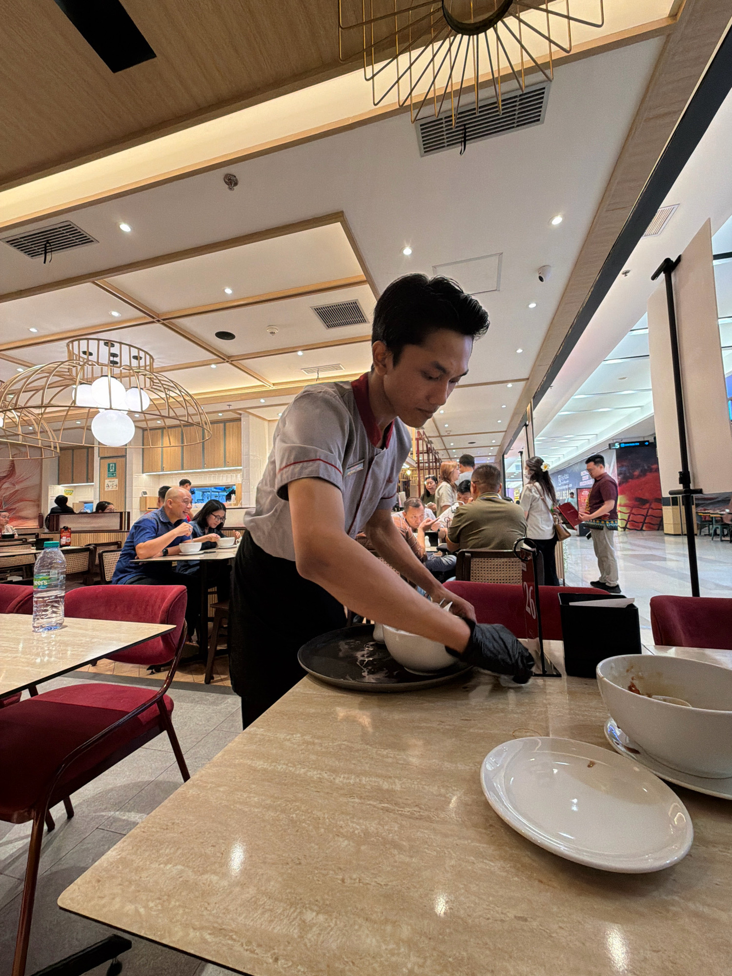 In a brightly lit, indoor dining establishment, a male server wearing a grey and red uniform and black gloves is seen from the waist up, focusing on cleaning a white bowl with a white cloth. He is standing behind a table with a beige, marbled surface, where a white plate and a large white bowl are visible. The server is in the foreground, slightly to the right of the image's center, and his hands are actively engaged in the cleaning process.
The background reveals other diners seated at tables, some engaged in conversation and eating. Several tables are covered in similar beige marbled surfaces, and red upholstered chairs are present. In the middle ground, a man in a blue shirt is smiling and looking at a woman in front of him, who is also looking at a bowl. Further back, more patrons are seated. The ceiling features modern lighting fixtures and air conditioning vents. The overall environment suggests a busy, contemporary restaurant or food court.