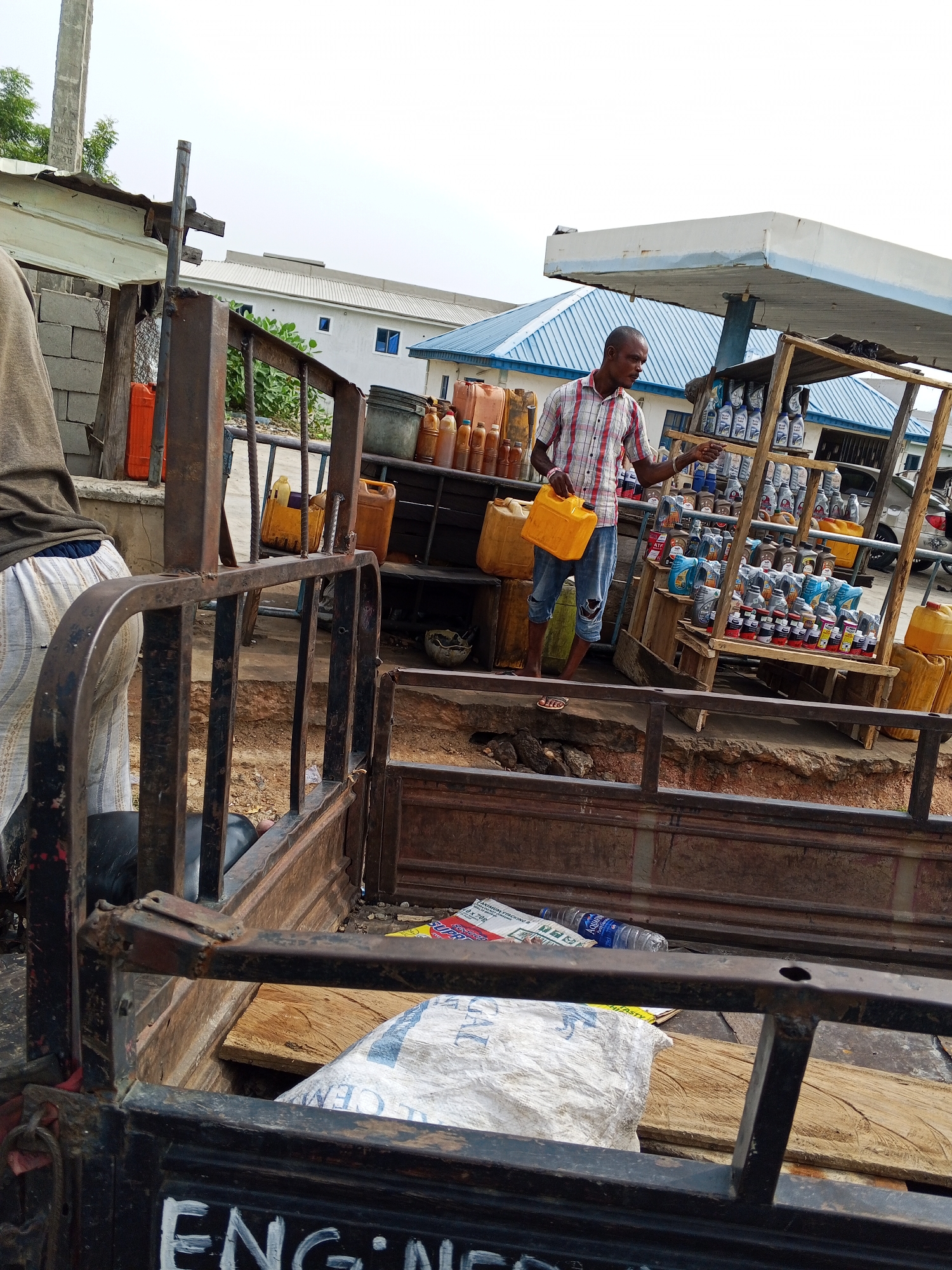The scene is located in Zango-ndaji, Nigeria. A man stands in front of a makeshift wooden stand displaying rows of bottled liquids, likely motor oil, in a roadside market or informal retail setting. He is wearing a checkered shirt, blue jean shorts, and sandals, holding a yellow plastic jerrycan. Behind him, a truck contains larger containers, possibly filled with fluids. The foreground reveals the rusty metal frame and bed of a vehicle, presumably used for transport, with scattered debris, including a white sack and water bottles. In the background is a building with a blue and white canopy, potentially a more established business or gas station. The overall impression is of an informal economic activity, suggesting a roadside market or trade in automotive supplies.