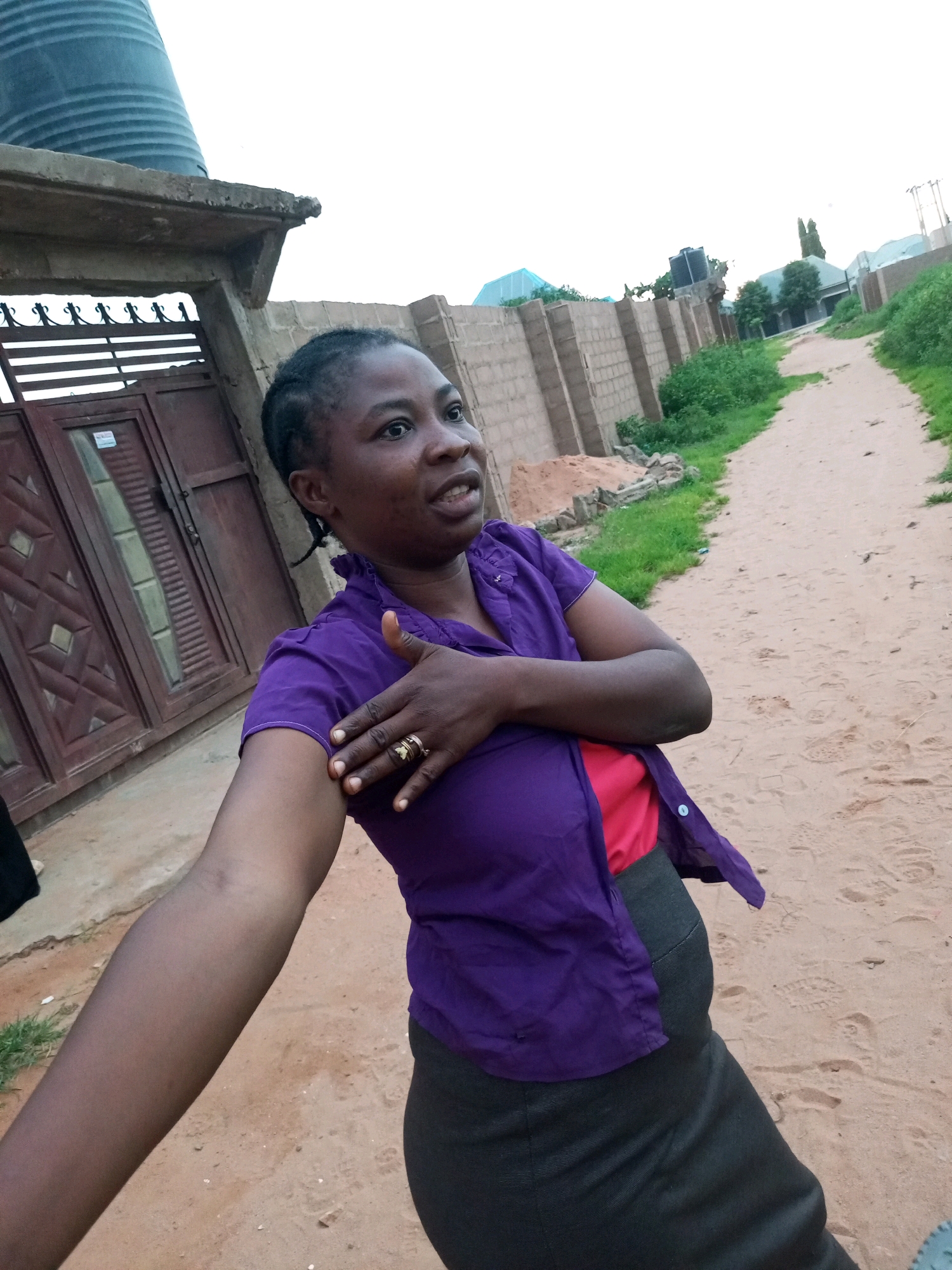 A medium shot captures a dark-skinned woman standing outdoors in what appears to be a dusty, unpaved area. She is looking to her right, with her mouth slightly open as if speaking. Her left arm is extended towards the camera, while her right hand is placed over her chest, touching the collar of her purple shirt. The woman is wearing a vibrant purple collared shirt with the top two buttons undone, revealing a glimpse of a red garment underneath. Her lower body is clad in a dark, fitted skirt. A gold ring with a prominent setting adorns her right ring finger.
The background suggests a semi-urban or rural setting. To the left of the woman is a large, dark brown double gate with intricate metalwork at the top. Beyond the gate, a concrete structure with a large, dark cylindrical object on top is visible, likely a water tank. A low, light-colored brick wall runs horizontally across the mid-ground, separating the area from a dirt path that recedes into the distance. Green vegetation flanks the path and the wall. In the far distance, buildings with corrugated metal roofs and utility poles can be seen. The overall lighting suggests it is either early morning or late afternoon, with an overcast sky. The ground is covered in fine, reddish-brown sand with visible footprints.