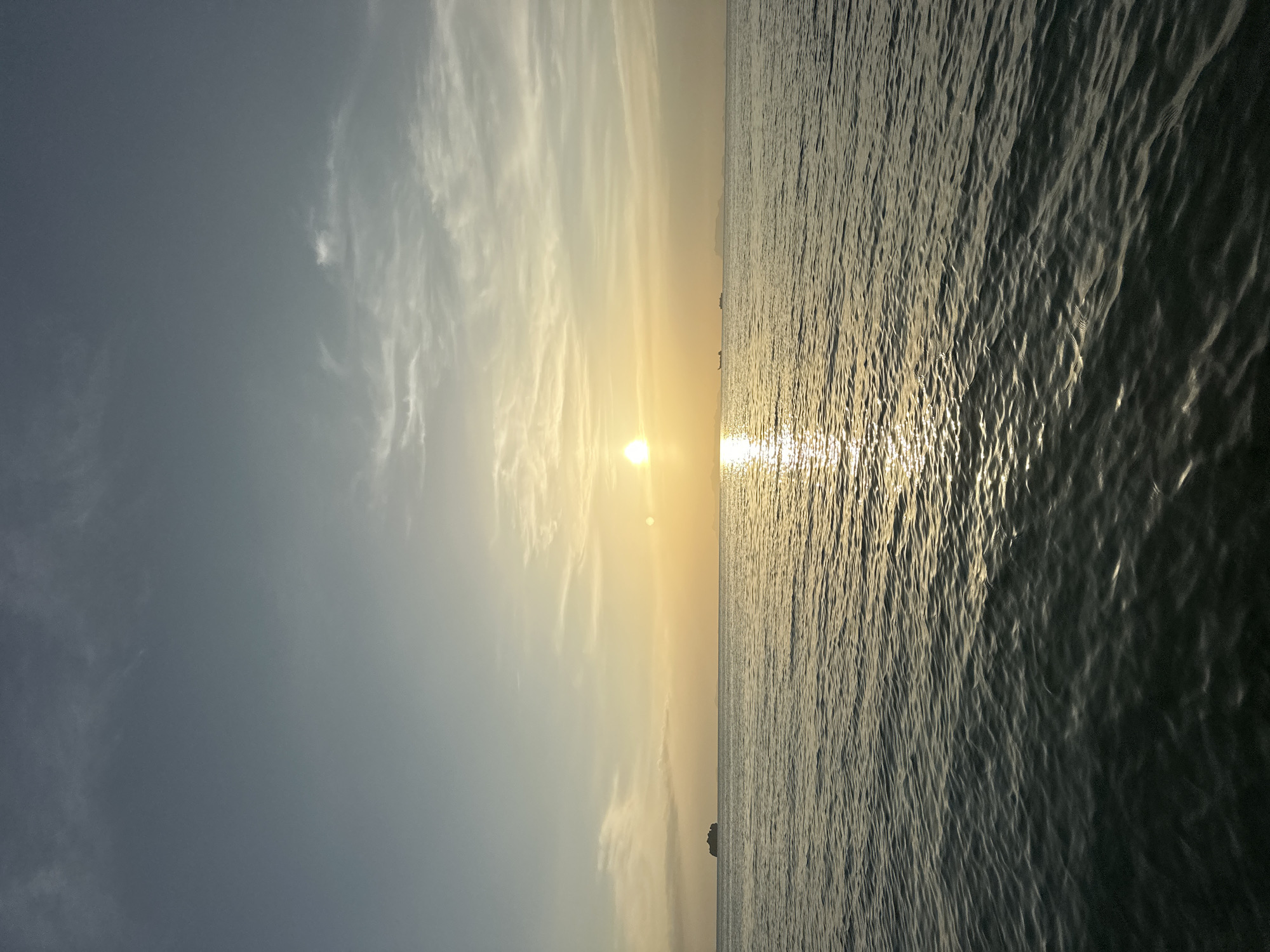 A serene sunset scene unfolds over the calm waters of the Gulf of Thailand, likely near Ko Samui District. The sun, a brilliant orb of light, hovers just above the horizon, casting a shimmering golden pathway across the gentle ripples of the sea. The sky transitions from a soft, pale blue at the top to warm hues of yellow and orange near the sun, with wispy white clouds scattered throughout. In the distance, the silhouettes of several vessels and a small island are visible, adding depth to the tranquil landscape. The foreground is dominated by the dark, undulating surface of the ocean, reflecting the warm tones of the sunset.