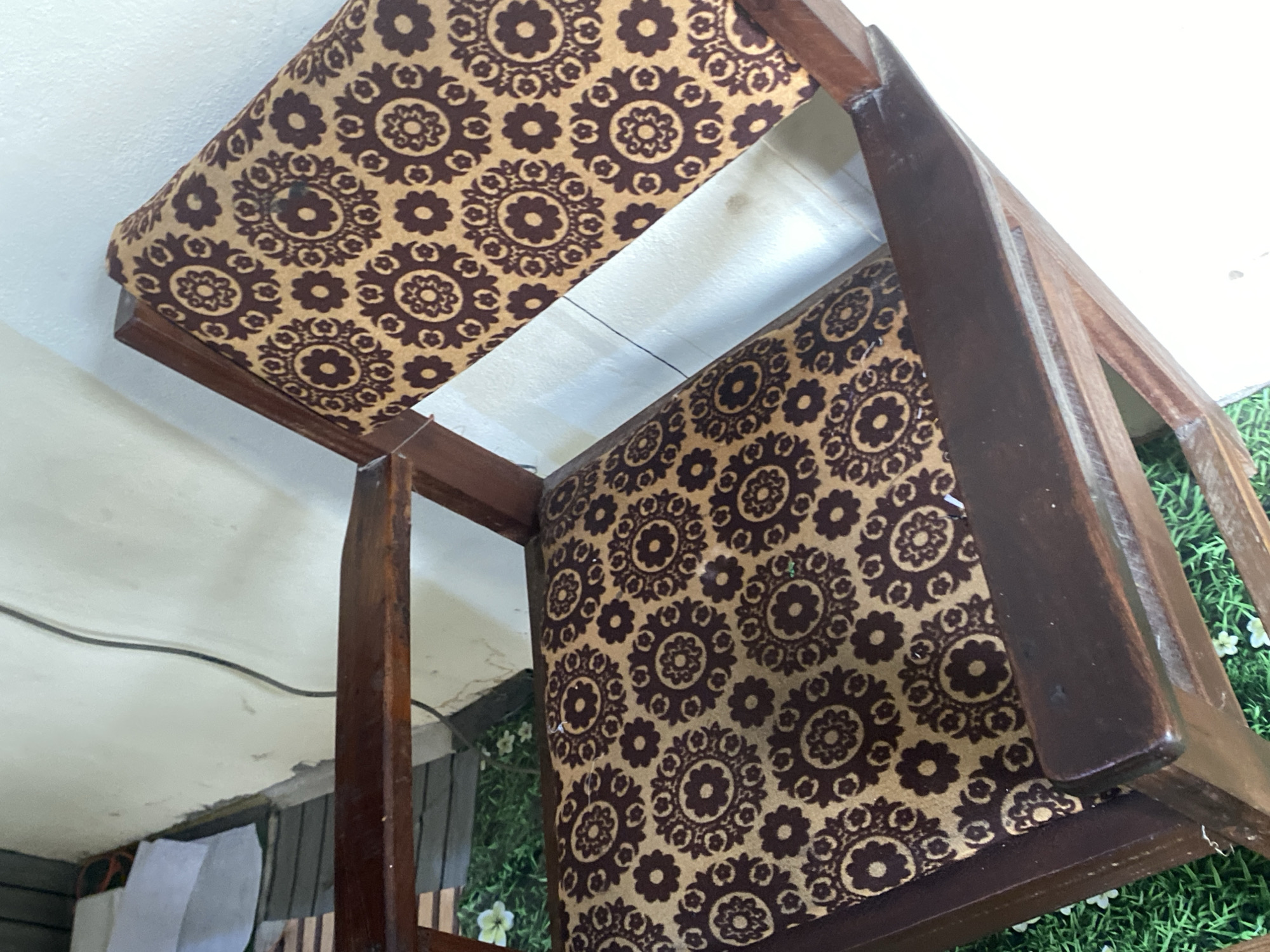 The image shows a close-up of an armchair with a floral pattern upholstery, situated indoors. The chair has a dark wooden frame. The upholstery fabric is a light tan color with a repeating pattern of dark brown circular motifs, each containing smaller petal-like shapes. The chair appears to be in good condition. The background is out of focus, but suggests a wall and possibly some objects on the floor. The context of Jauro Yinu, Nigeria is not evident from the visual information in the image.