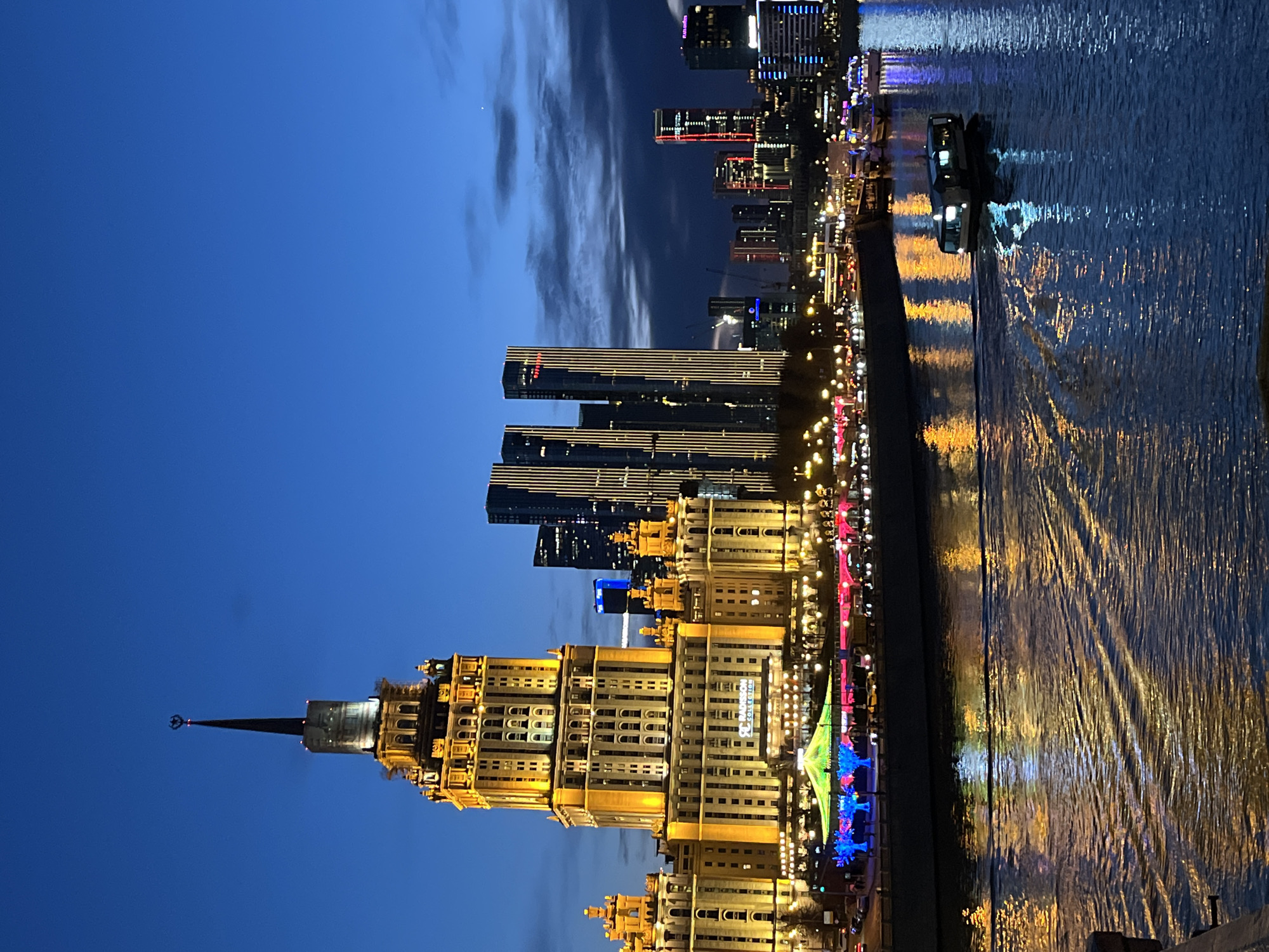 The image depicts a night view of Moscow, Russia. The Radisson Collection Hotel is prominently featured in the foreground, with its distinctive Stalinist architecture illuminated in warm yellow light. Several modern skyscrapers are visible behind the hotel, also lit up against the dark blue sky. The Moscow River flows along the right side of the frame, reflecting the city lights and adding to the vibrant atmosphere. A boat cruises on the river. The sky is a deep blue with wisps of cloud. Streetlights line the embankment.