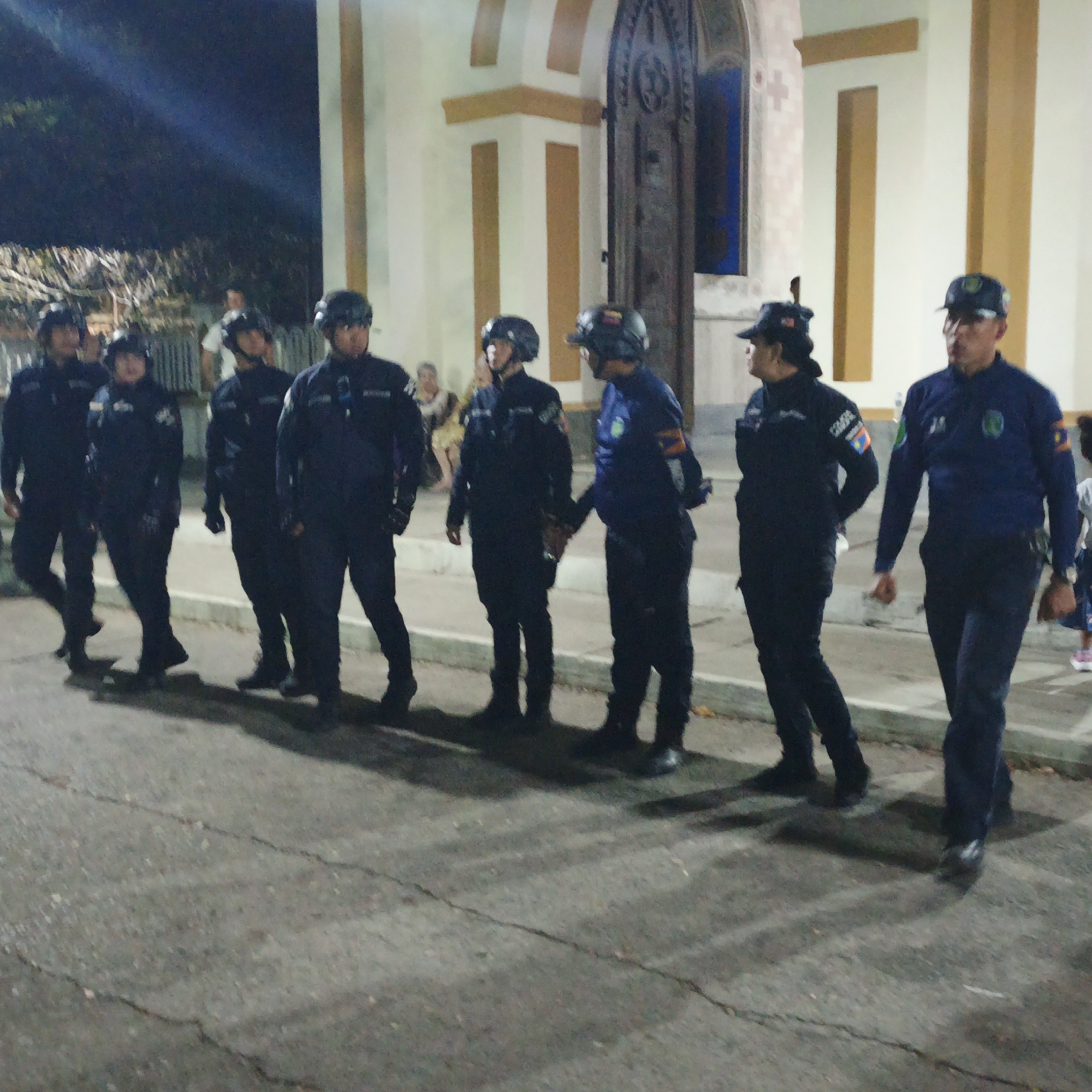 A group of at least eight individuals, likely law enforcement officers due to their dark uniforms and some wearing helmets, are standing or walking on a paved surface in front of a building. The building has a white facade with yellow trim. A large door or entrance with arched details is visible. The time appears to be night or dusk, with artificial lighting present. This image was taken in Tinaquillo, Venezuela.