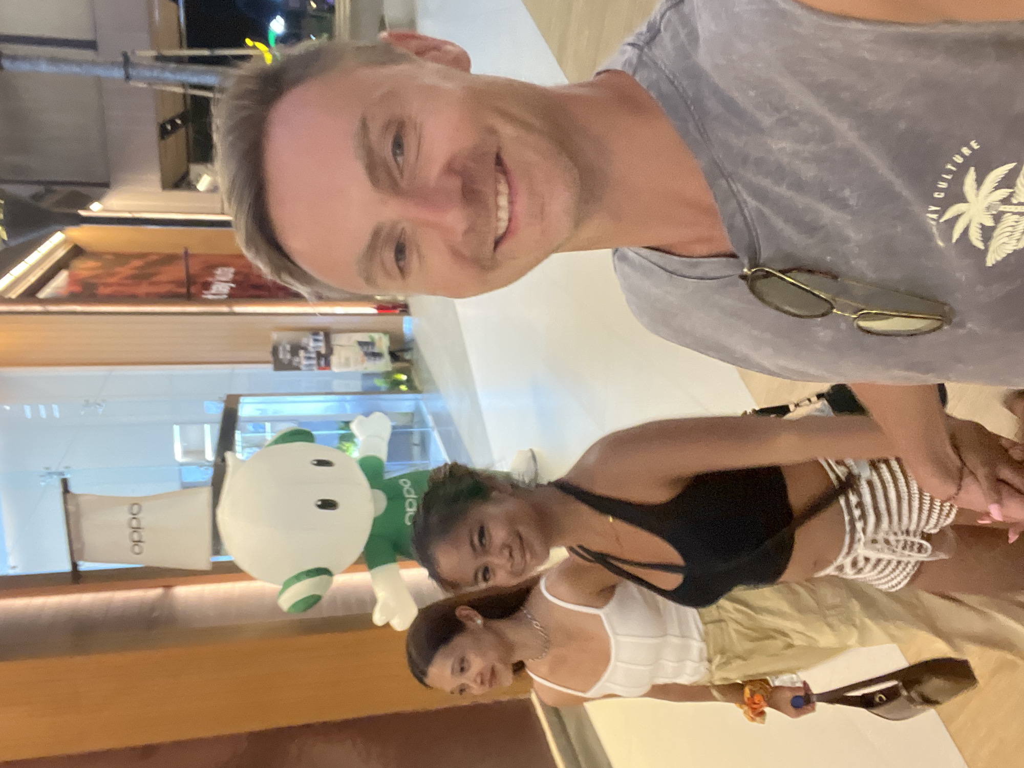 The image is a selfie taken in what appears to be a shopping mall in Kuta, Indonesia. The photographer is a smiling white male wearing a grey t-shirt and sunglasses. Two young women of Asian descent are in the background. One wears a white top, the other a black top with patterned shorts, and is holding a purse. An "OPPO" sign and an oversized, green and white mascot are visible in the background, confirming it's likely an OPPO store or promotional display in the mall. The lighting suggests it is evening or indoor light. The background of the image shows retail storefronts and mall ambiance.