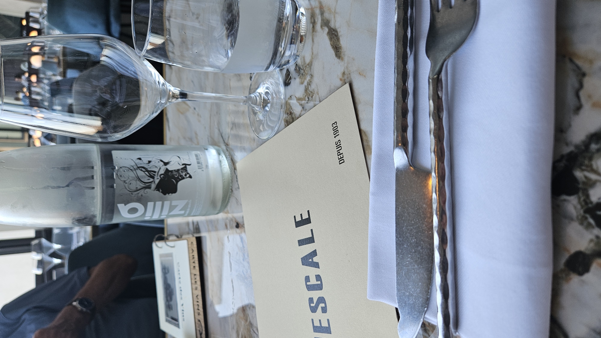 The image shows a close-up view of a table setting at a restaurant. A bottle of Zilia water, a wine glass, and a water glass are visible, along with a menu that reads "VESCALE DEPUIS 1903". A knife and fork are laid out on a white napkin. In the background, a person wearing a blue shirt and a watch can be seen. The restaurant appears to be located in the city of L'Île-Rousse, France, as indicated by the menu's inscription. The table has a marble-like surface, and the overall ambiance suggests a formal dining setting.