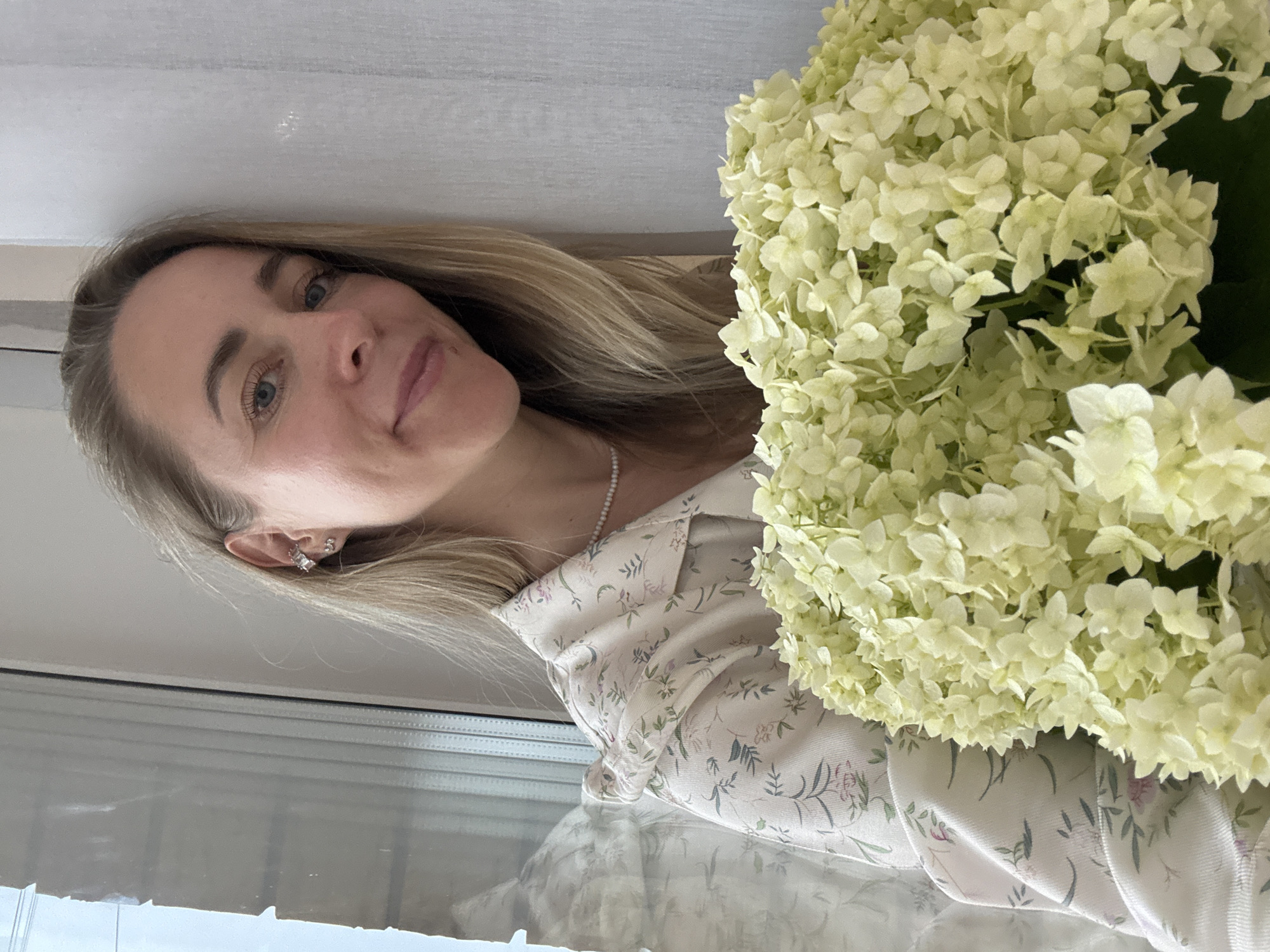 A woman with blonde hair and blue eyes is captured in a close-up portrait. She is wearing a light-colored blouse with a floral pattern and a pearl necklace. Her left ear is adorned with delicate earrings. She is holding a large bouquet of pale yellow hydrangeas, which partially obscures her body. The woman appears to be looking slightly off-camera with a soft smile.
The setting appears to be indoors, with a window or reflective surface visible in the background, suggesting a home or studio environment. The lighting is soft and diffused, possibly indicating daylight. The overall mood is serene and elegant, with a touch of natural beauty provided by the flowers. The image is oriented vertically, with the woman's head towards the top. There is no visible text in the image. The provided location context of Chaikovsky, Russia, does not offer specific visual cues within the image itself.
