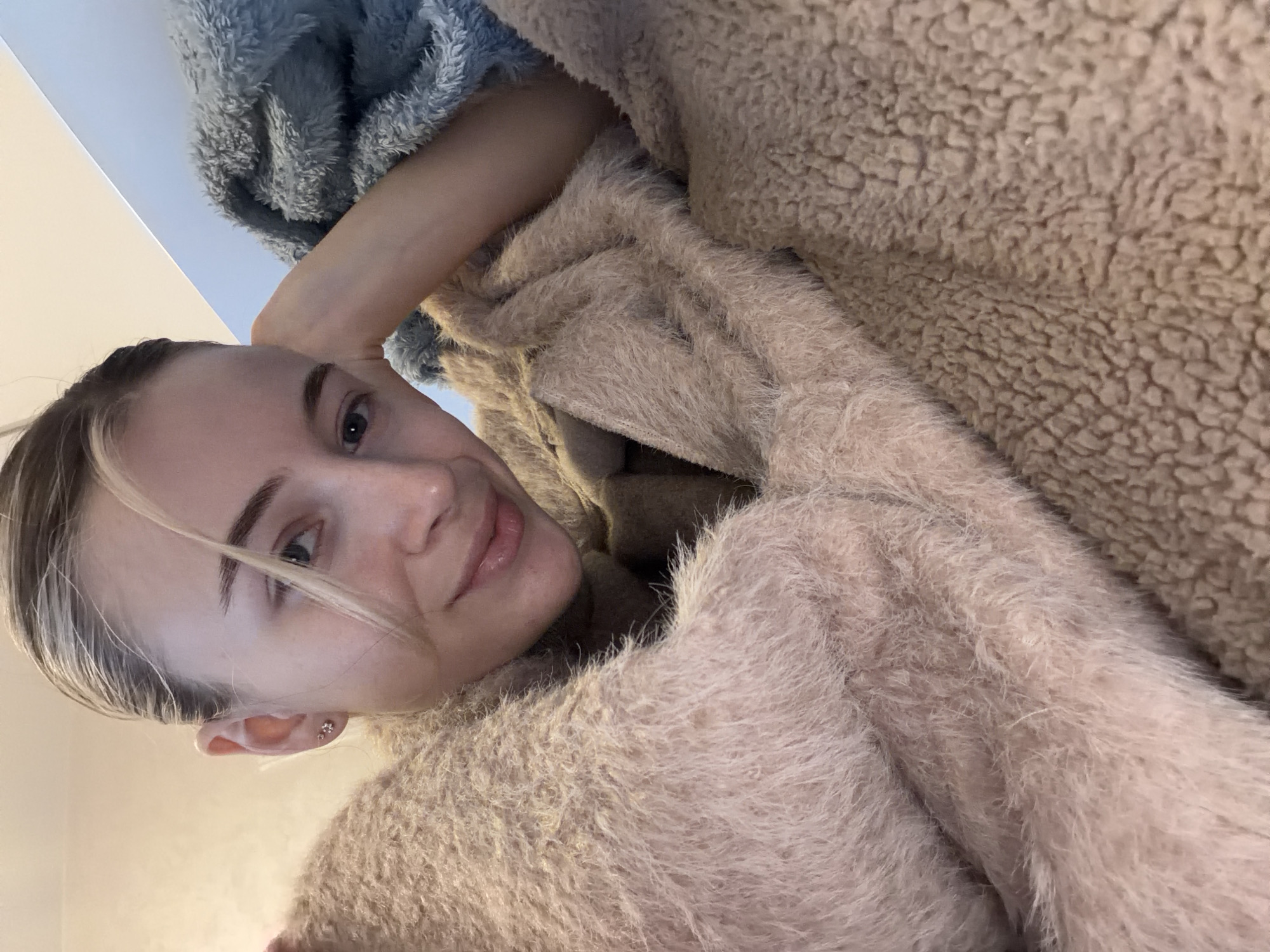 A woman with blonde hair pulled back is looking at the camera. She is wearing a fuzzy, light pink or beige sweater. Her left arm is raised and bent behind her head. She is lying against a fluffy, tan blanket and a fluffy, light blue blanket. The location is not discernible in the image.