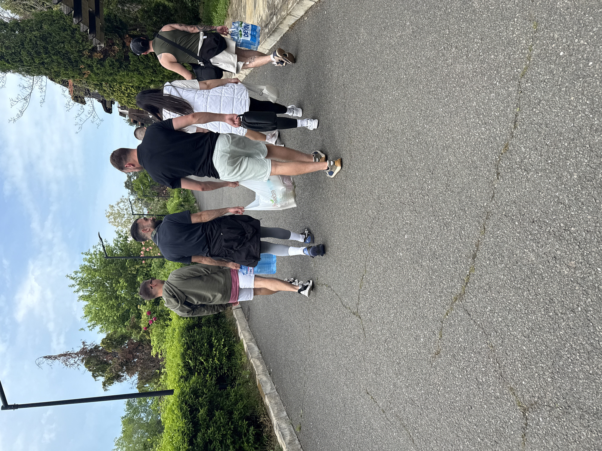 A group of five individuals walks single file on an asphalt path in Sozopol, Bulgaria. The group moves from left to right. The path is bordered by greenery on the left, including shrubs, trees, and vegetation. A man in a green jacket and pink shorts leads, followed by a man in black with gray tights, a man in black with white shorts carrying a Fresko bag, a woman in a white jacket and black pants, and a man in green with a black bag and tattoos. The last two people carry bottles of Devin water. The sky is visible in the background with some clouds. A black lamp post is visible near the greenery. Some buildings are visible behind the trees.