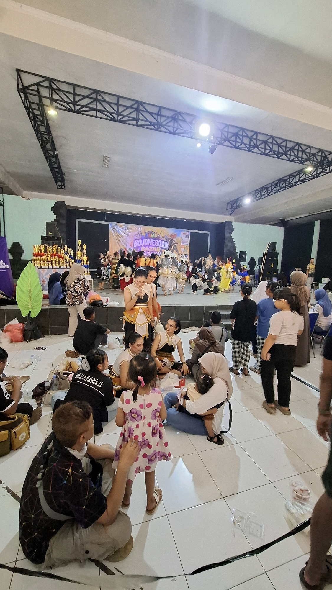 The image depicts an indoor event in Bojonegoro, Indonesia, likely a cultural performance or competition, given the stage setup and the presence of trophies. The scene is a medium shot, capturing a diverse group of people, both on a raised stage and seated on the floor.
On the stage, several individuals are dressed in traditional costumes, suggesting a performance. Some are standing, while others are seated, possibly awaiting their turn or resting. The backdrop of the stage features a banner with the text "BOJONEGORO" prominently displayed, along with other decorative elements. Trophies are arranged on shelves to the left of the stage.
In the foreground and midground, many people are seated on the tiled floor, indicating an audience or participants. They are a mix of adults and children. Some individuals are interacting, such as a woman holding a baby and another woman with a young girl in a floral dress. There are also bags and other personal items scattered on the floor.
The environment appears to be a large indoor space with a high ceiling supported by black metal trusses. Lighting fixtures are visible on the trusses. The overall atmosphere suggests a community gathering or event in Bojonegoro.