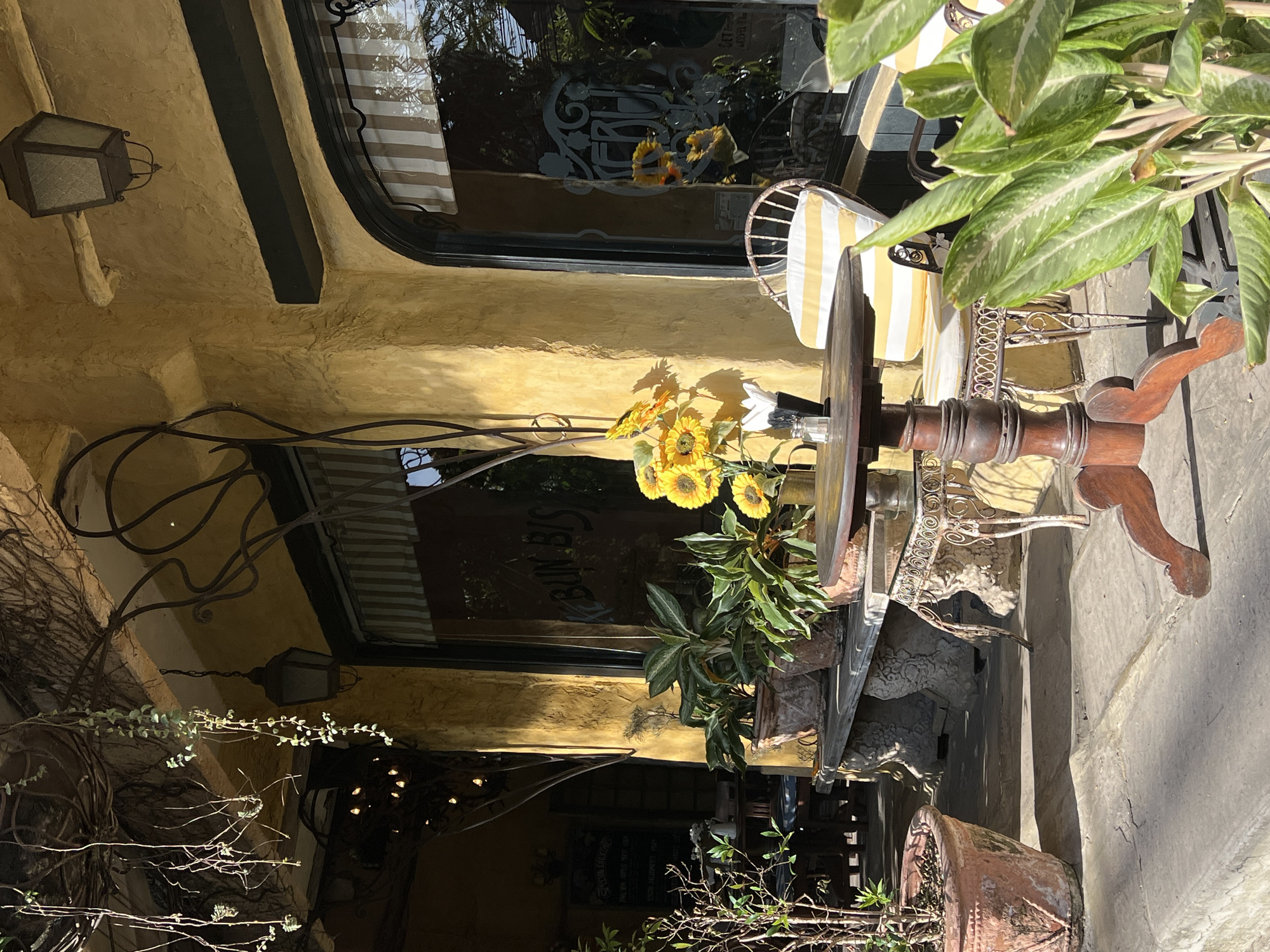 The image shows an outdoor seating area in what appears to be a commercial district. The setting is the Ubud District in Indonesia. Visible architectural elements include a textured, yellow-painted facade with black trim, window awnings with beige and white stripes, and wrought iron details. A table, adorned with a vase of sunflowers and a water bottle, is the focal point. The table is accompanied by a chair with a yellow and white striped cushion. Potted plants are abundant, adding to the tropical atmosphere. A hanging lantern is affixed to the facade. The overall scene suggests a café or restaurant. Reflections of an emblem reading "BUN BIS" are on the window.