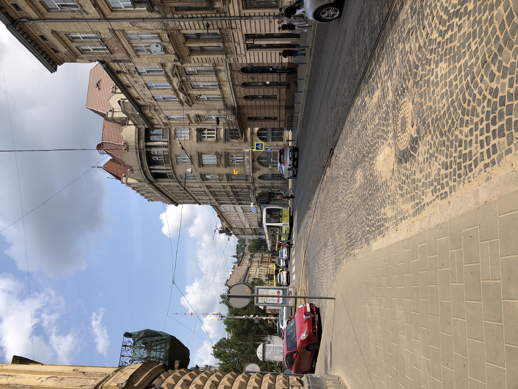 The image captures a street scene in Lviv, Ukraine, likely in the historic center, given the architecture and cobblestone streets. The perspective is from a sidewalk looking down a street lined with old, ornate buildings. The buildings exhibit classical architectural styles with detailed facades, balconies, and red-tiled roofs.
In the foreground, a red Alfa Romeo hatchback is parked on the left side of the street, adjacent to a sidewalk paved with rectangular gray tiles. The street itself is paved with dark cobblestones and features tram tracks.
Further down the street, a modern yellow and white tram is visible, stopped or moving slowly. Several cars are also present on the street, including a silver hatchback driving away from the camera and other vehicles further in the distance.
A few people are scattered along the sidewalk and crossing the street, appearing to be pedestrians. The sky is partly cloudy with a mix of blue sky and gray clouds. The overall impression is that of a lively, historic European city street on a bright day.