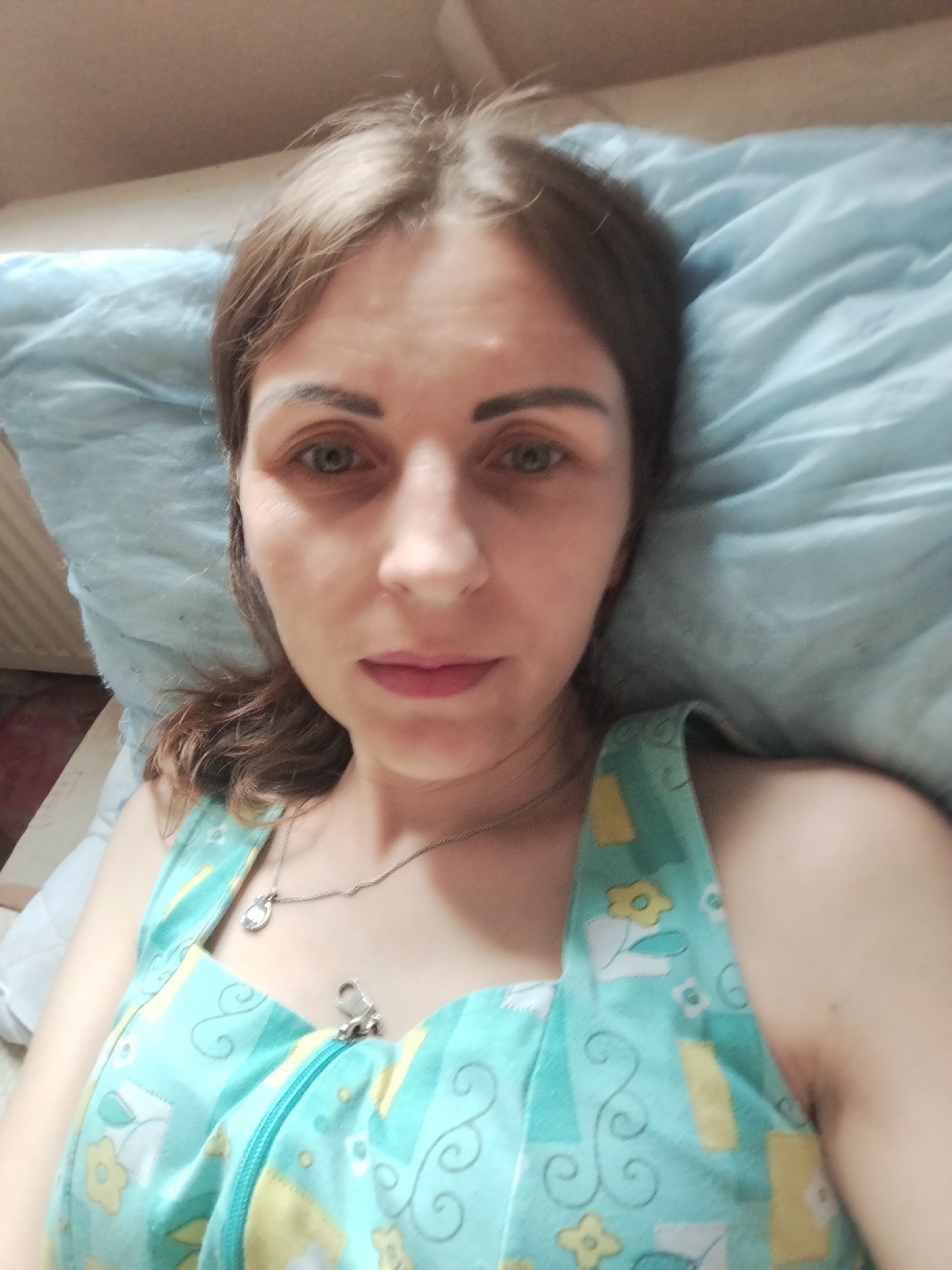 A woman is lying in bed, resting her head on a light blue pillow. She is looking directly at the camera. The woman has light brown hair, blue eyes, and is wearing a light blue nightgown with a floral pattern. A silver necklace with a pendant is visible around her neck. The room appears to be a bedroom, with a wall visible in the background. The lighting suggests it is daytime. The image was taken in Vinnytsia, Ukraine.