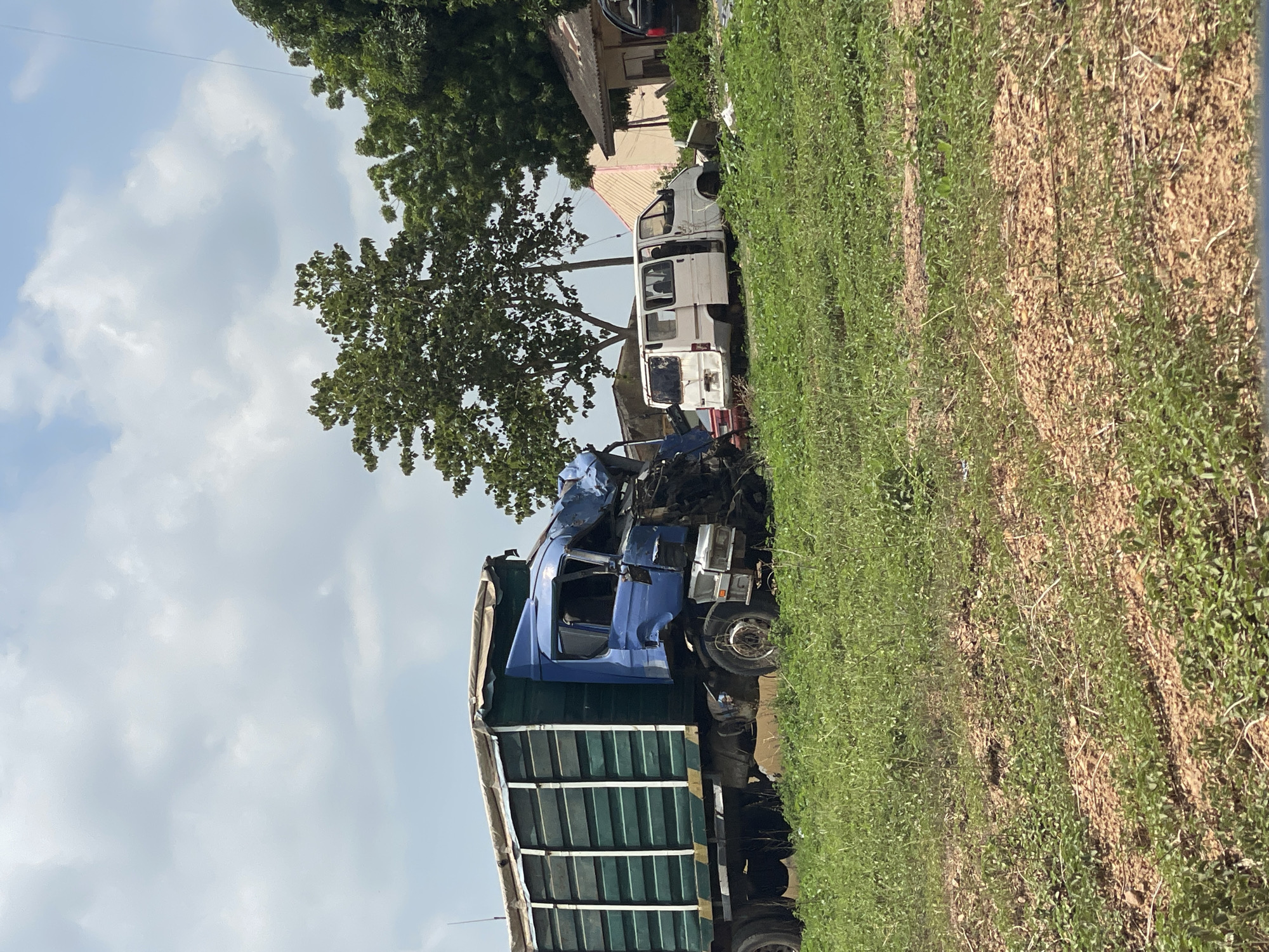 The image depicts the aftermath of a vehicular collision in an area near Akwanga, Nigeria. A blue cargo truck with a green-slatted trailer has sustained significant front-end damage, including a crushed cabin. A white van is also nearby, partially obscured, with visible damage. The crash occurred on a roadside area with vegetation. A tree stands near the vehicles. The sky is cloudy.