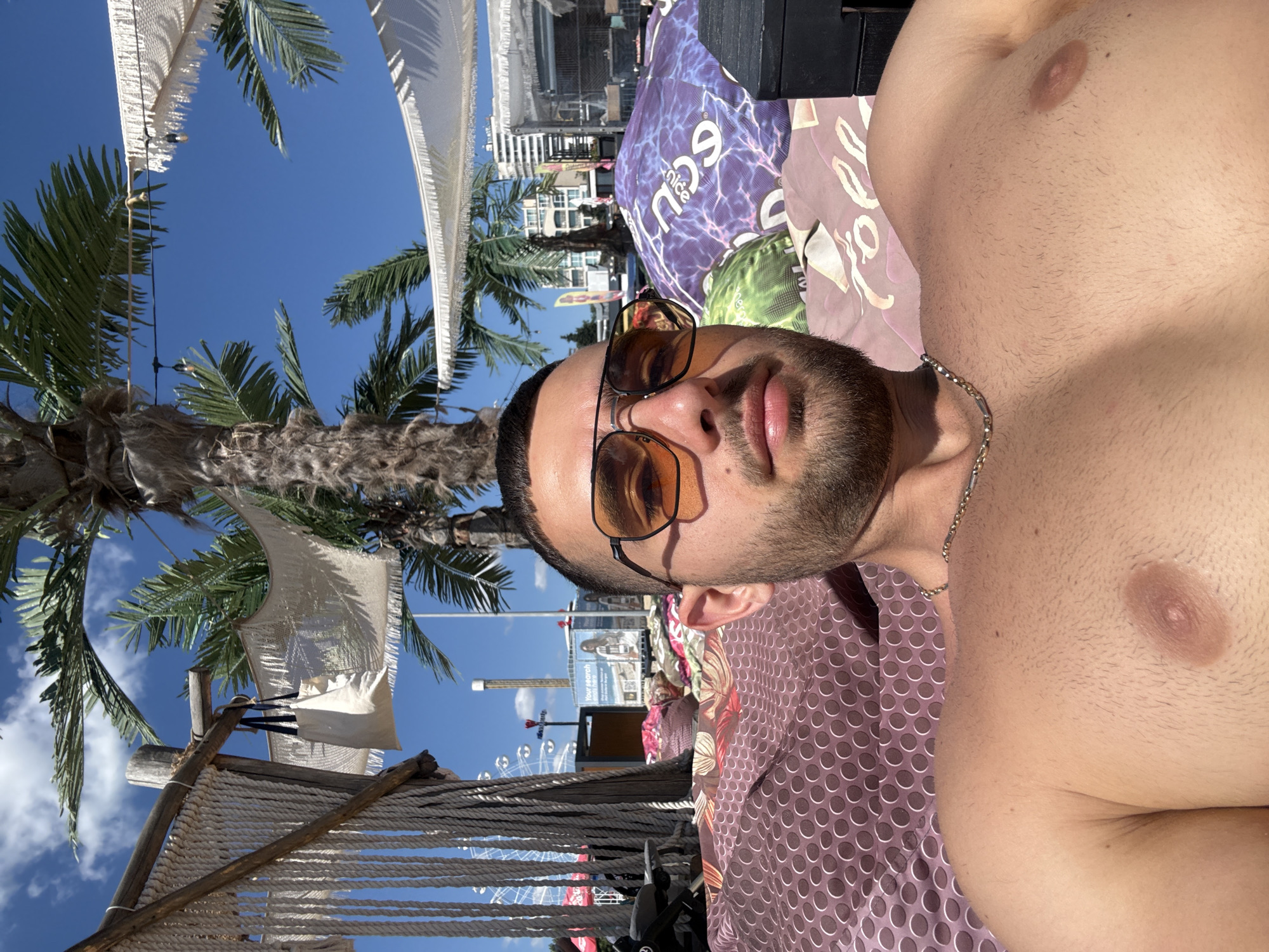 A man, with dark hair and a beard, wearing aviator sunglasses, is lying on a patterned lounger outdoors. He is shirtless and has a gold chain around his neck. Behind him, palm trees and a clear blue sky are visible, suggesting a beach or resort setting. In the background, a Ferris wheel and buildings can be seen, indicating an urban or tourist area. The overall scene appears to be a sunny day at a recreational location in Nesebar, Bulgaria.